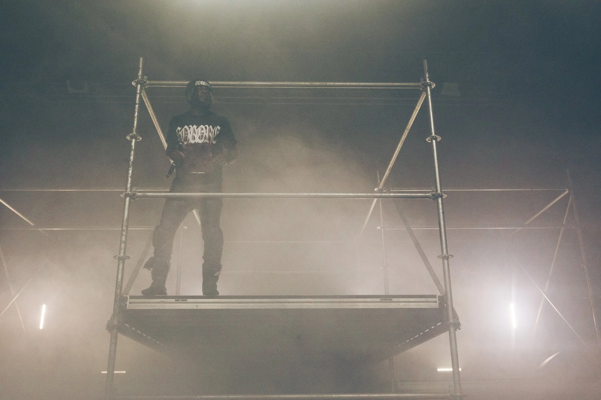 A person standing on a metal scaffolding platform in a foggy or smoky environment, wearing a black outfit and a headband with white lettering.