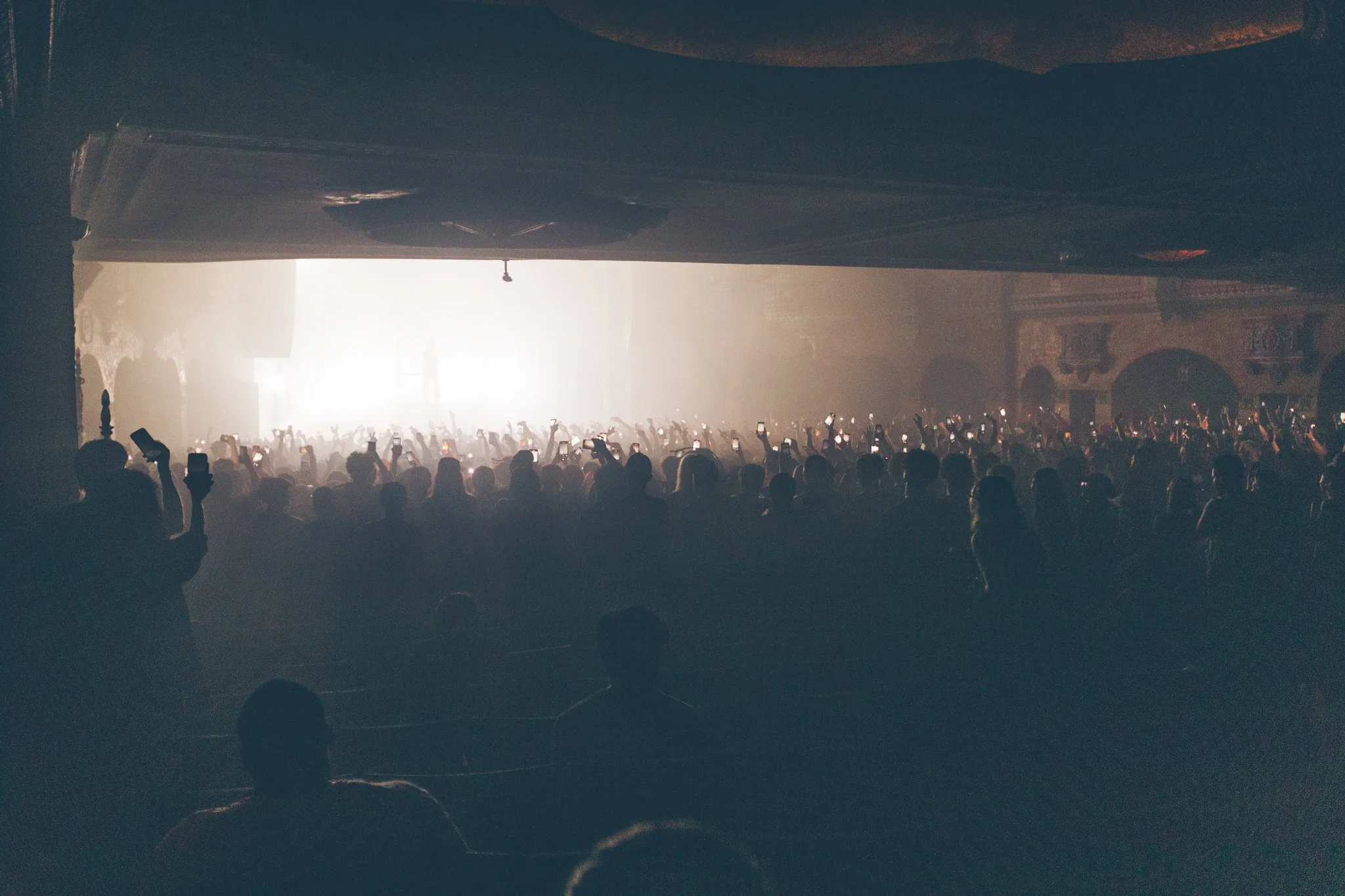 Crowd at concert or event with many people holding up phones, illuminated by stage or screen lighting in a large hall or theater.