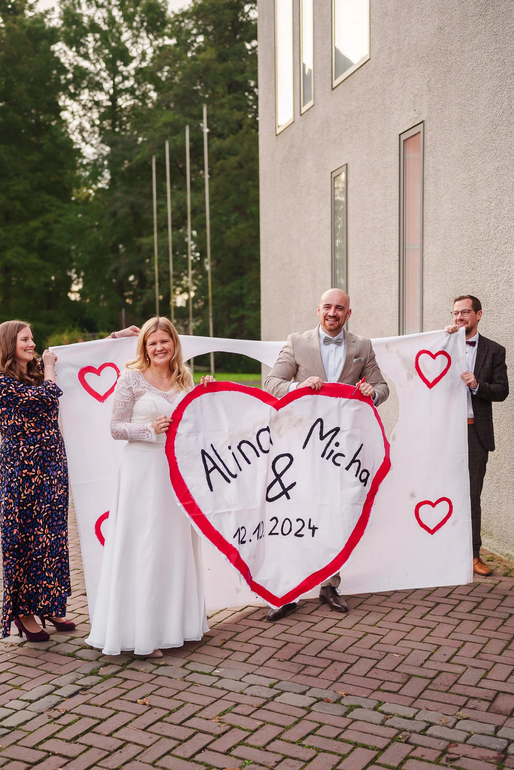 Gruppe von fünf Menschen bei einer Hochzeit, die ein Banner mit den Namen Alina und Micha und dem Hochzeitsdatum 12.10.2024 halten, draußen vor einem modernen Gebäude.