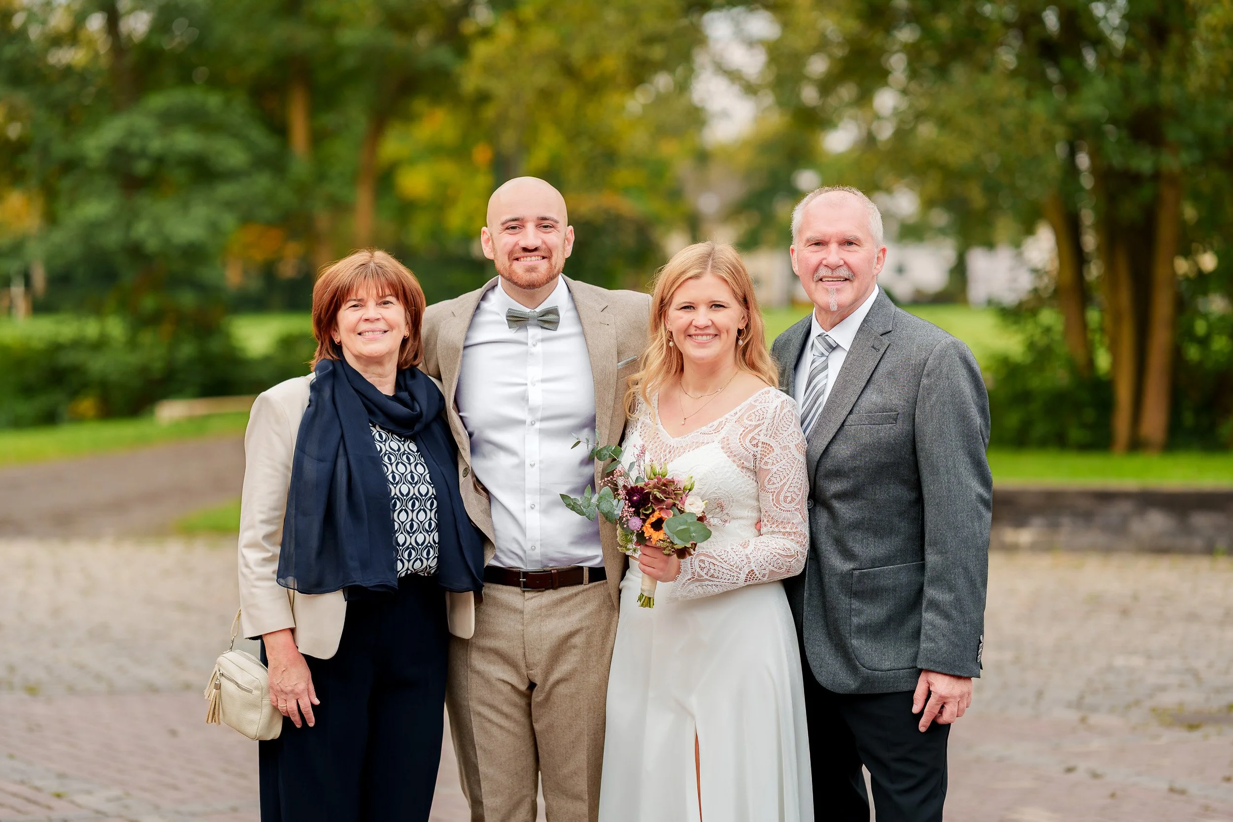 Ein glückliches Brautpaar mit zwei älteren Menschen bei einer Hochzeit im Freien, umgeben von grünen Bäumen.