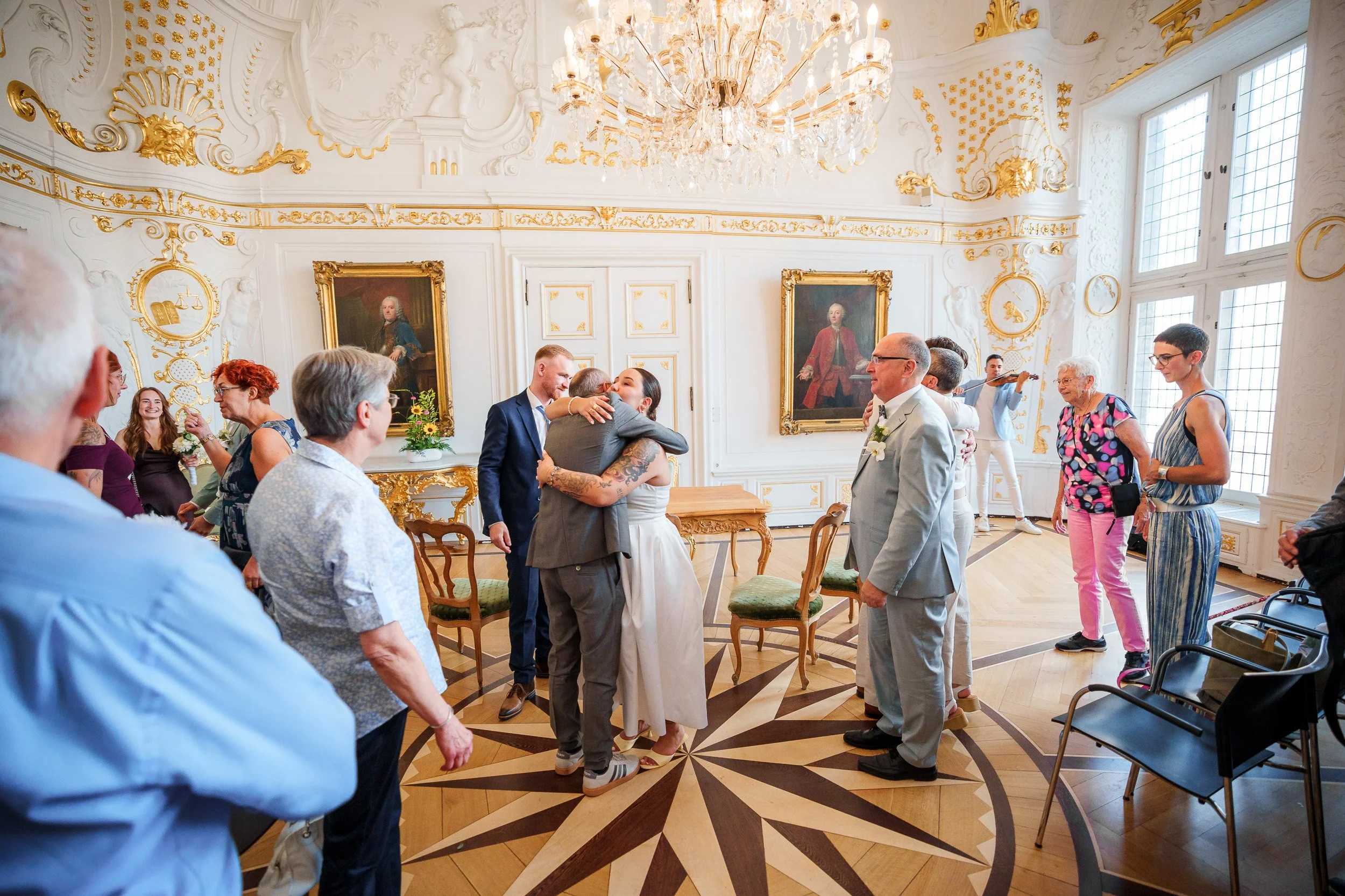 Eine Gruppe von Leuten in einem eleganten, mit Gold verzierten Raum, einige umarmen sich, während ein Musiker im Hintergrund spielt.