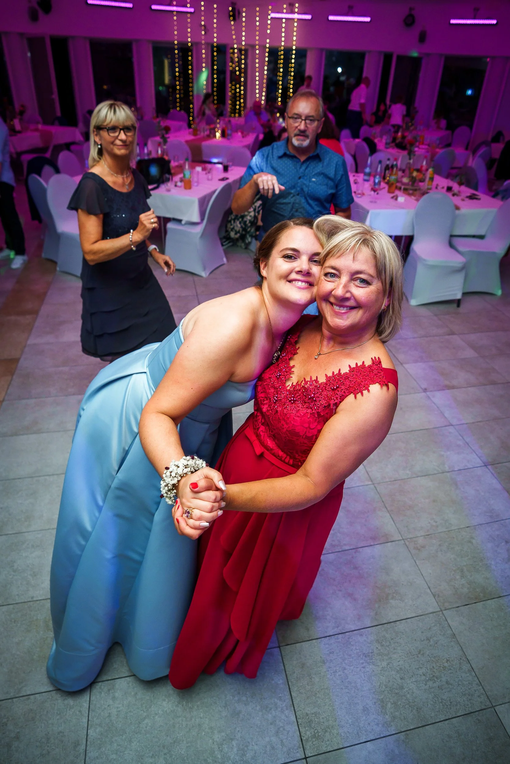 Zwei Frauen tanzen bei einer Feier, umarmt und lächelnd, im festlich dekorierten Raum mit Tischen und Gästen im Hintergrund.