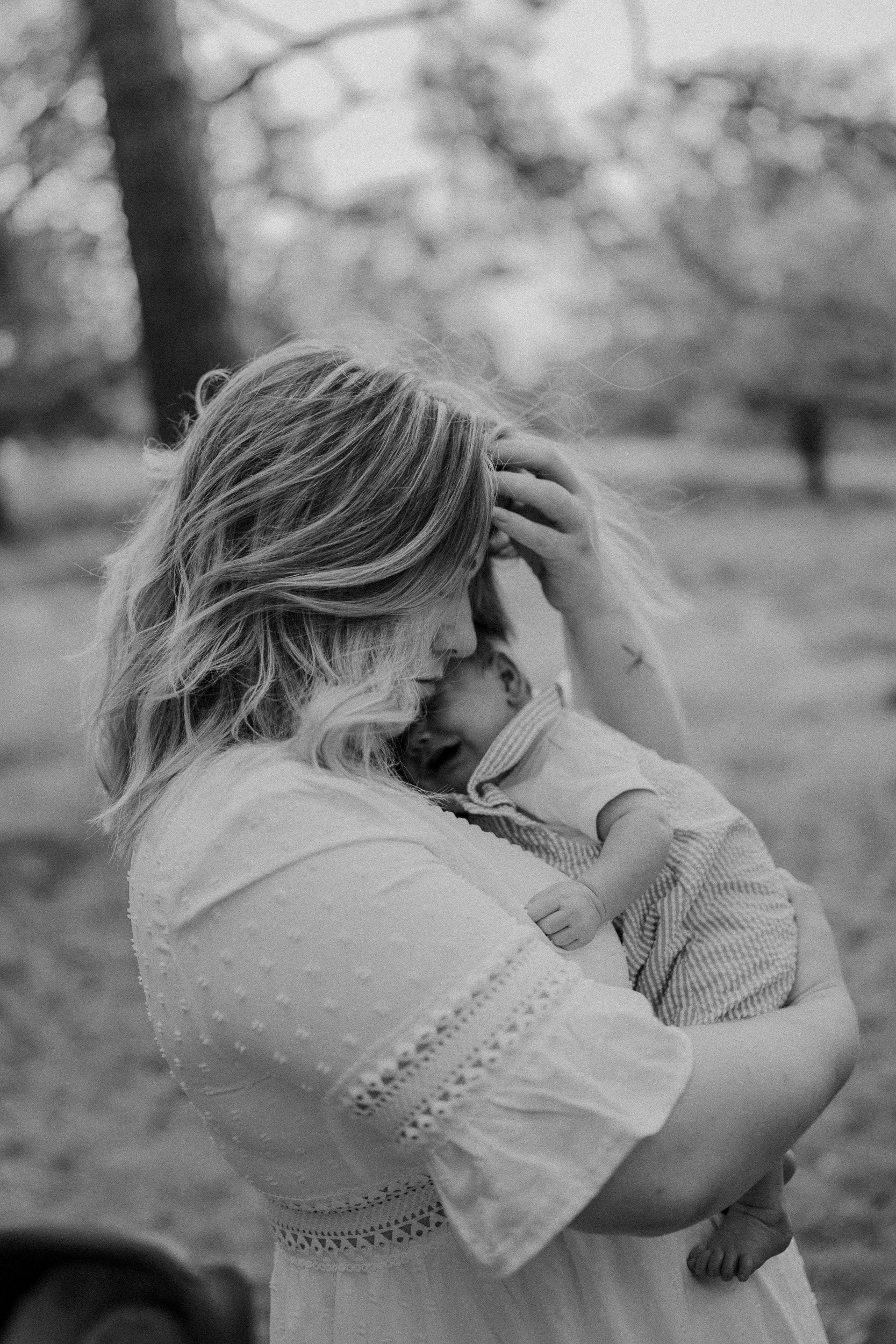 Eine Frau hält ein weinendes Baby in den Armen, umgeben von Bäumen im Freien, in Schwarz-Weiß-Fotografie.