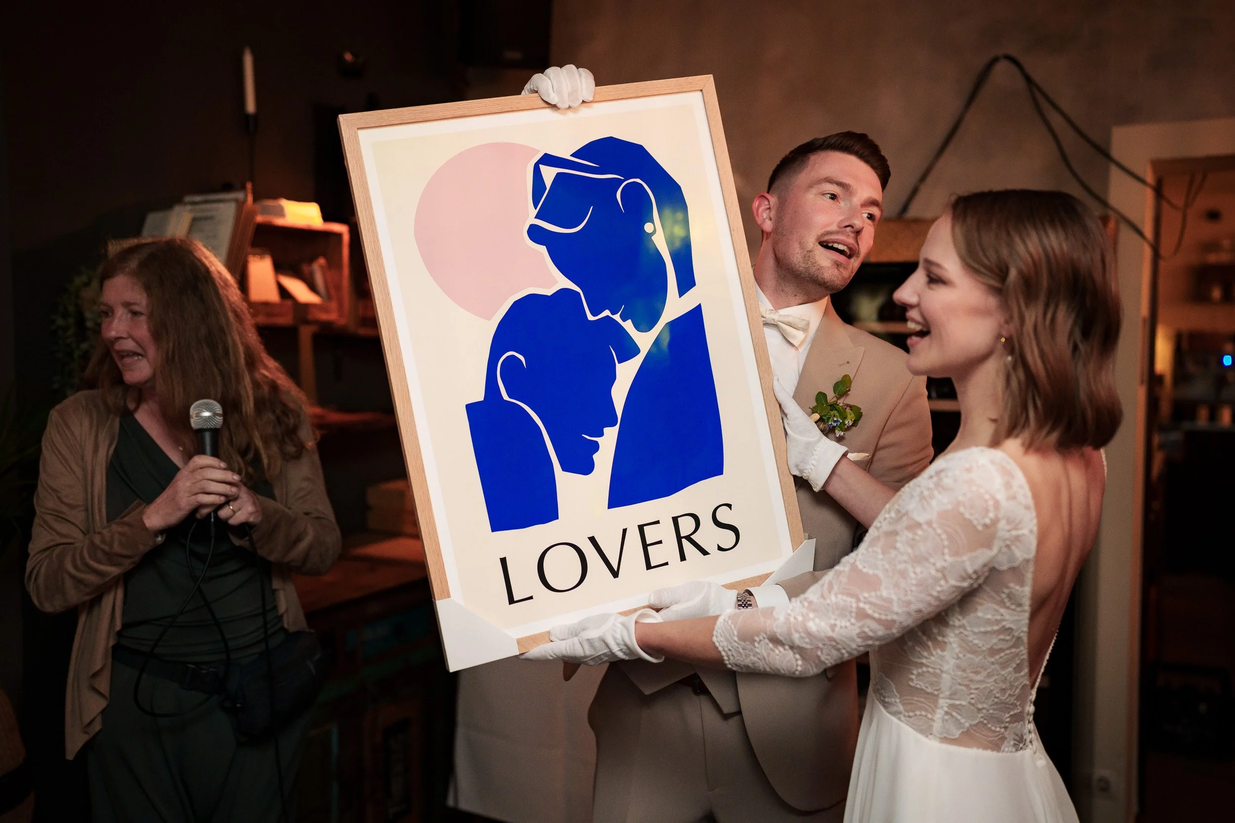 Ein Brautpaar bei einer Hochzeitsfeier, das ein Bild mit dem Wort 'LOVERS' und illustrativen Gesichtern hält, umgeben von Gästen.
