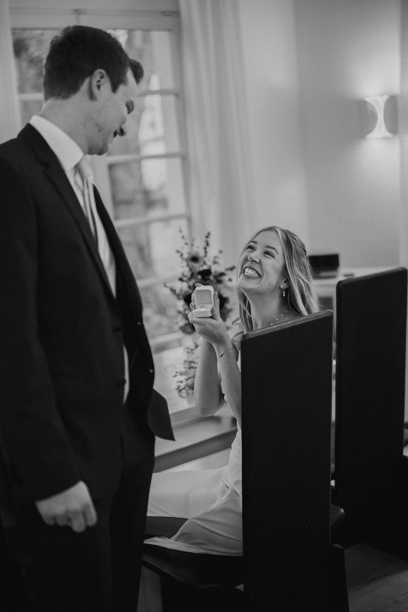 Ein lachendes junges Mädchen, das einen Ring in einer Geschenkbox hält, während ein junges Mann in einem Anzug auf sie blickt.