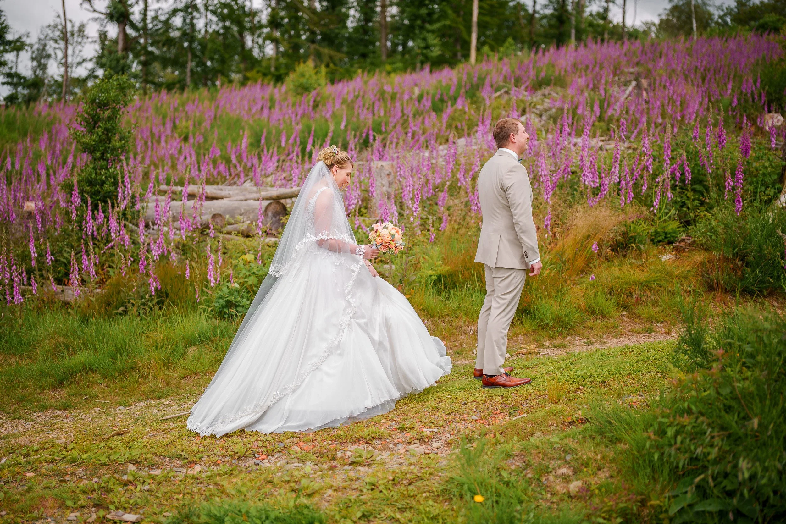 Ein Brautpaar auf einem Pfad in einem blühenden Garten mit lila und pink Farben, die Braut trägt ein weißes Hochzeitskleid mit Schleier, der Bräutigam trägt einen hellen Anzug.