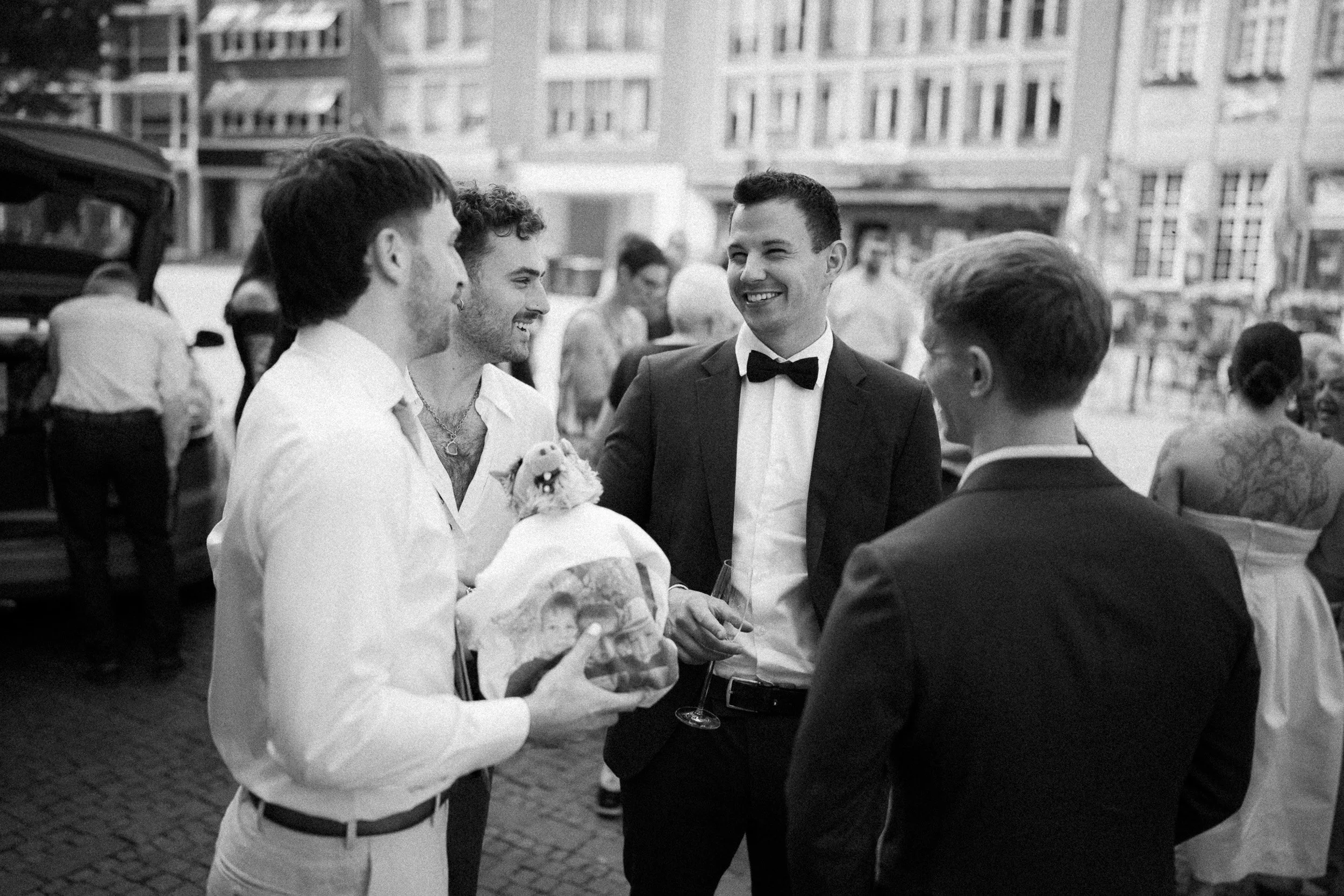 Eine Gruppe von Männern in formeller Kleidung, die auf einer Feier im Freien miteinander sprechen. Es scheint eine festliche Veranstaltung zu sein, möglicherweise eine Hochzeit.