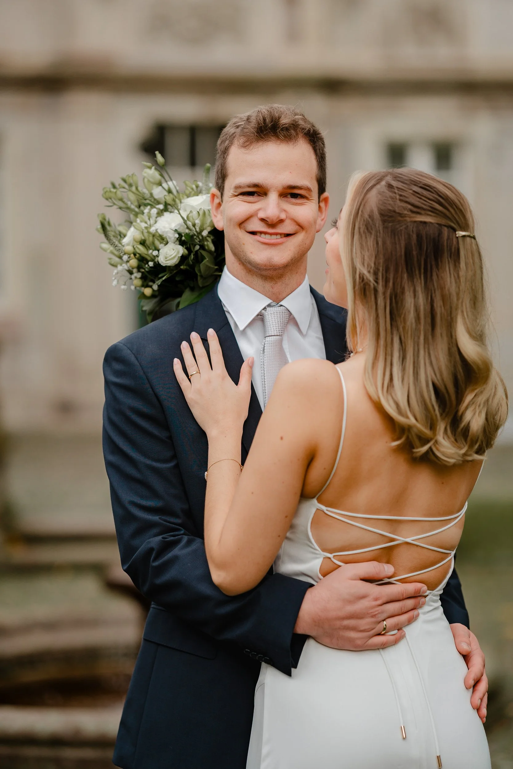 Ein Paar bei ihrer Hochzeit, der Mann in Anzug und die Frau in einem weißen Kleid, umarmend vor einem Gebäude im Freien.