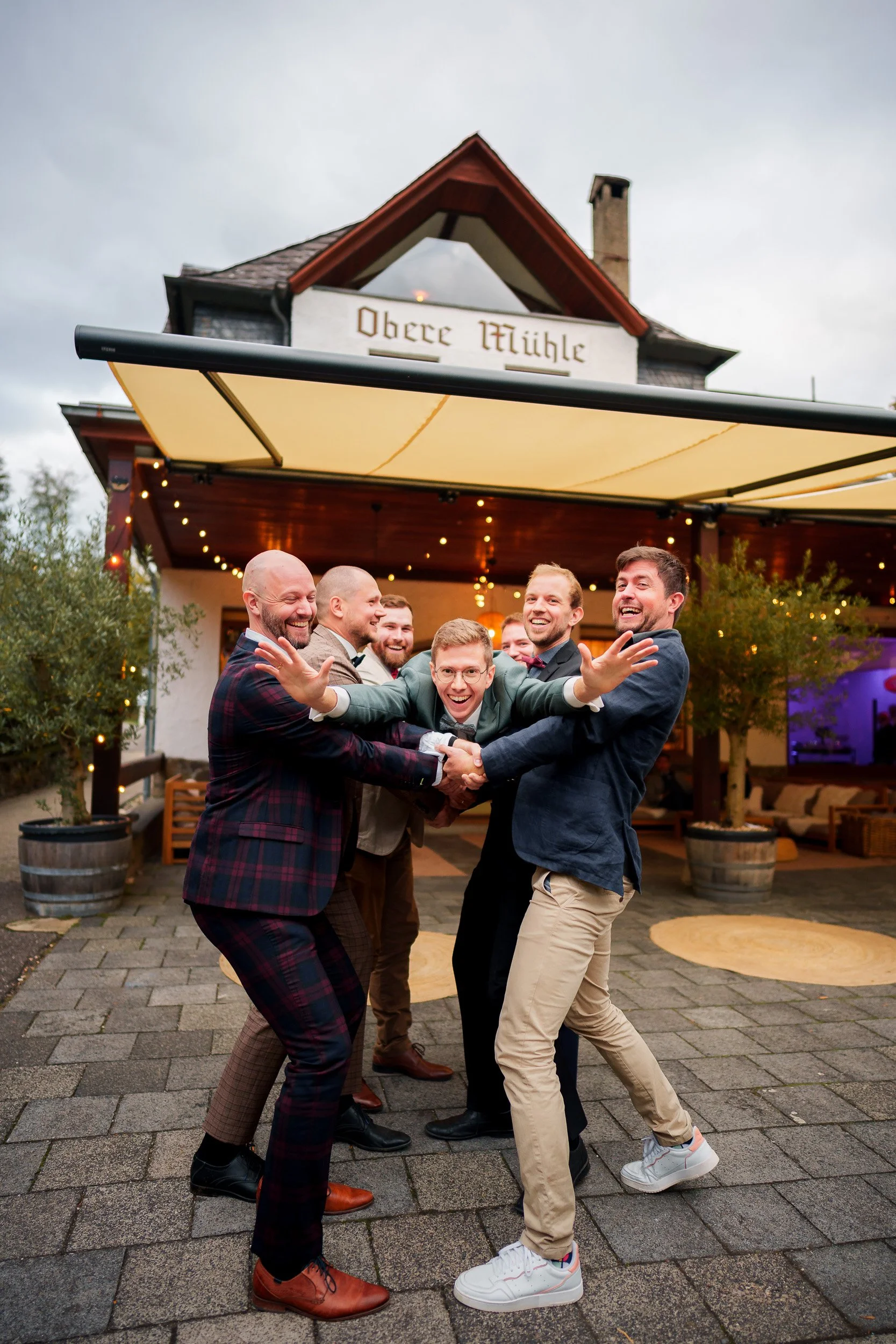 Gruppe von Männern, die sich bei einer Feier im Freien umarmen und Lauthals lachen, vor einem Gebäudeeingang mit der Aufschrift 'Obere Mühle' und einer hell erleuchteten, festlichen Dekoration.