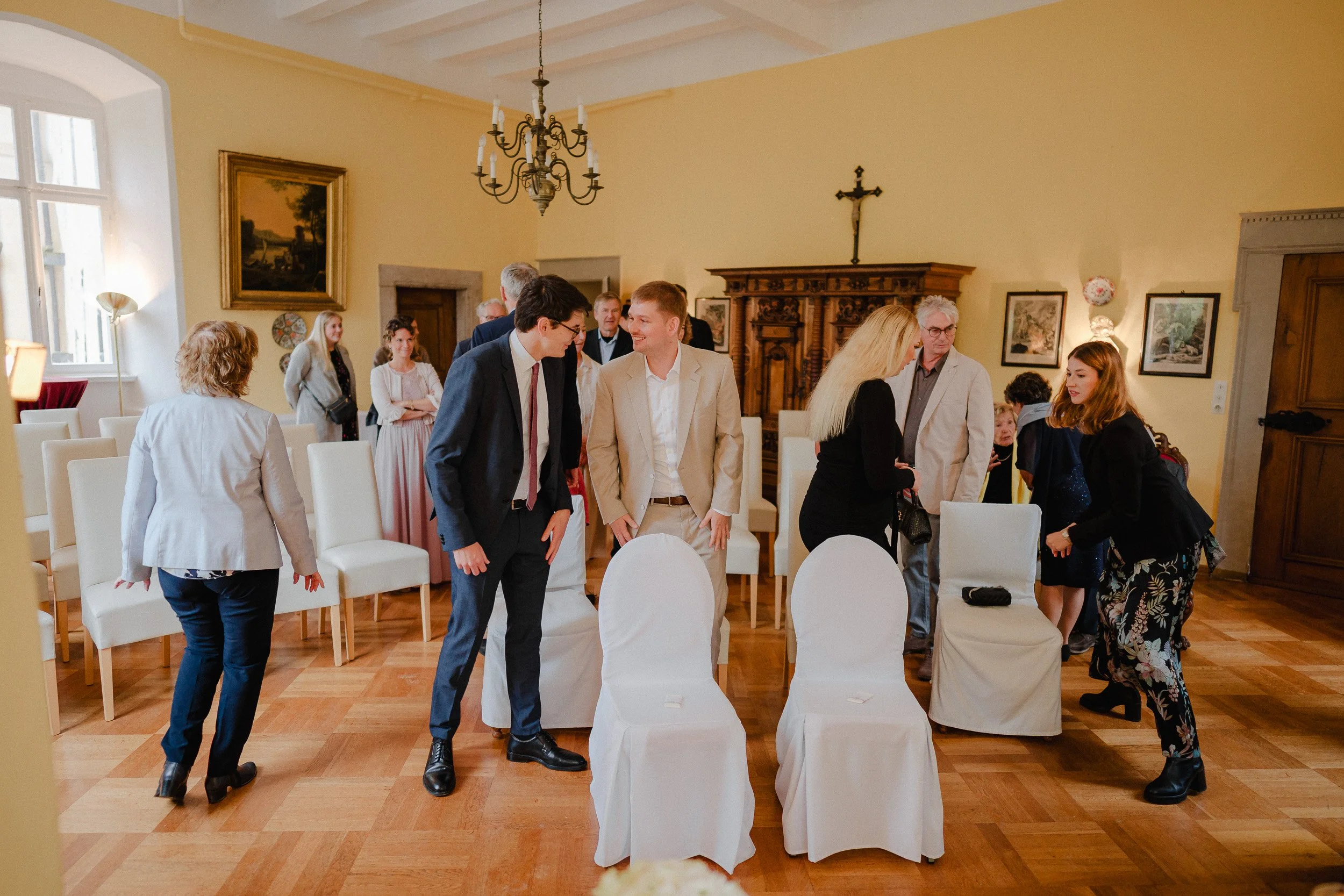 Menschen bei einer gesellschaftlichen Veranstaltung im Raum, einige sitzen, andere stehen und unterhalten sich, es gibt einen Kronleuchter an der Decke und Bilder an den Wänden.