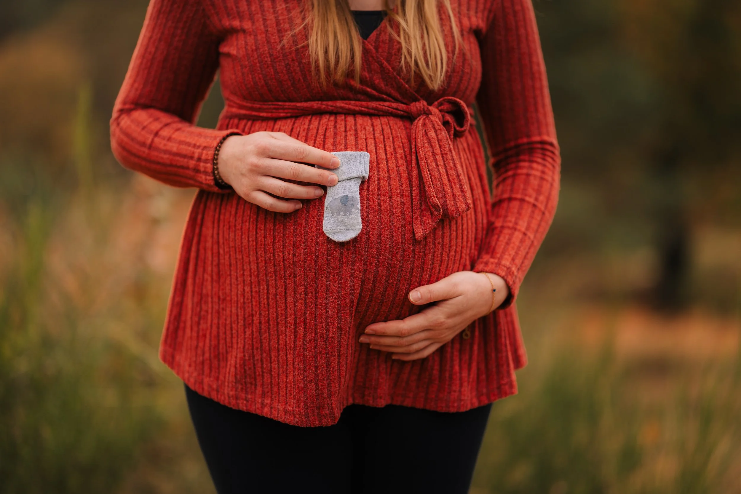 Schwangere Frau, die in der Natur im Herbst steht, trägt ein rotes Kleid, hält einen kleinen grauen Babystrampler mit Eulenmuster vor den Bauch.