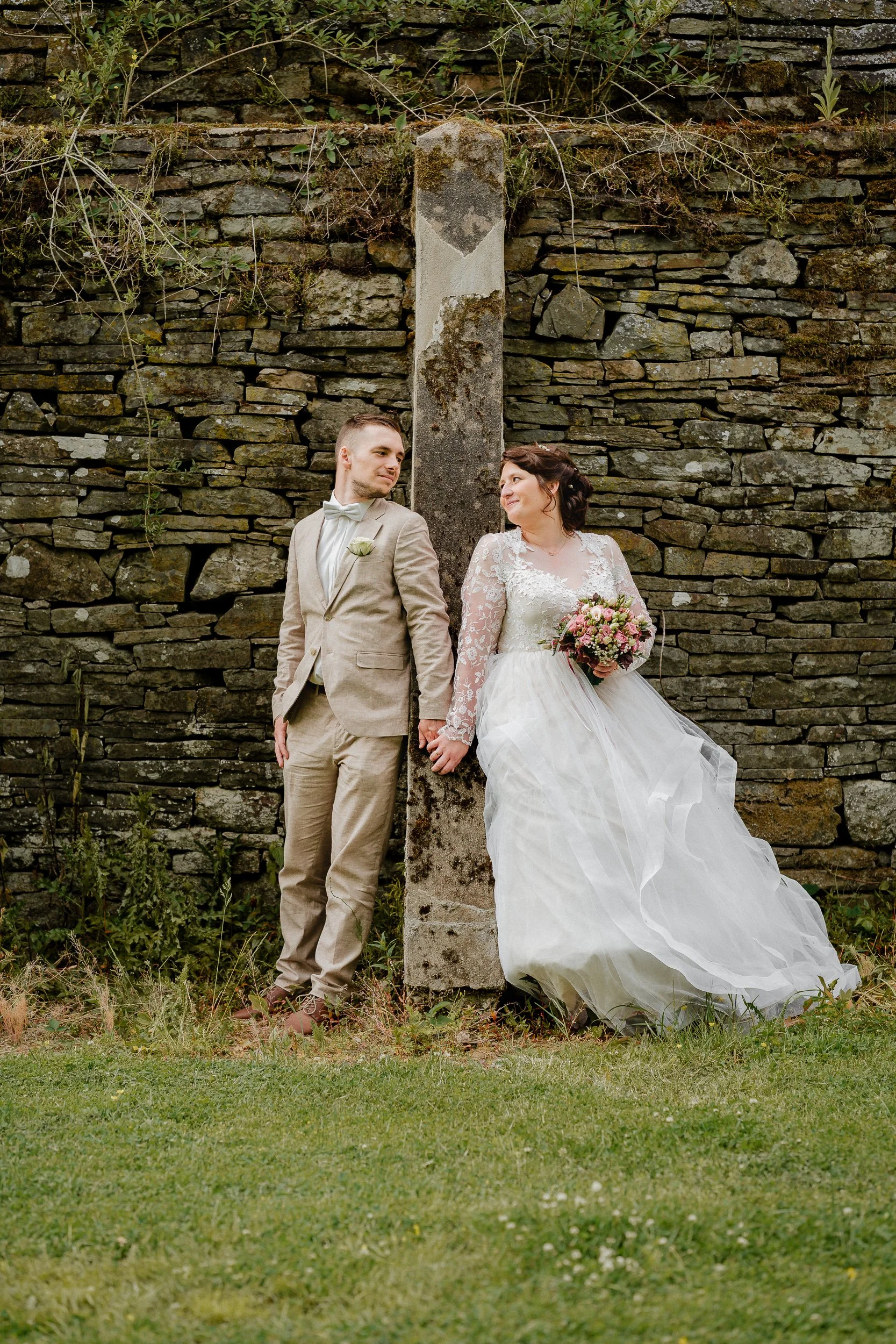 Ein frisch verheiratetes Paar steht an einem alten Steinmauer mit einer Säule in der Mitte. Der Bräutigam trägt einen hellen Anzug mit Fliege, die Braut ein weißes Brautkleid mit Spitze, hält einen Blumenstrauß, und sie schauen sich liebevoll an.