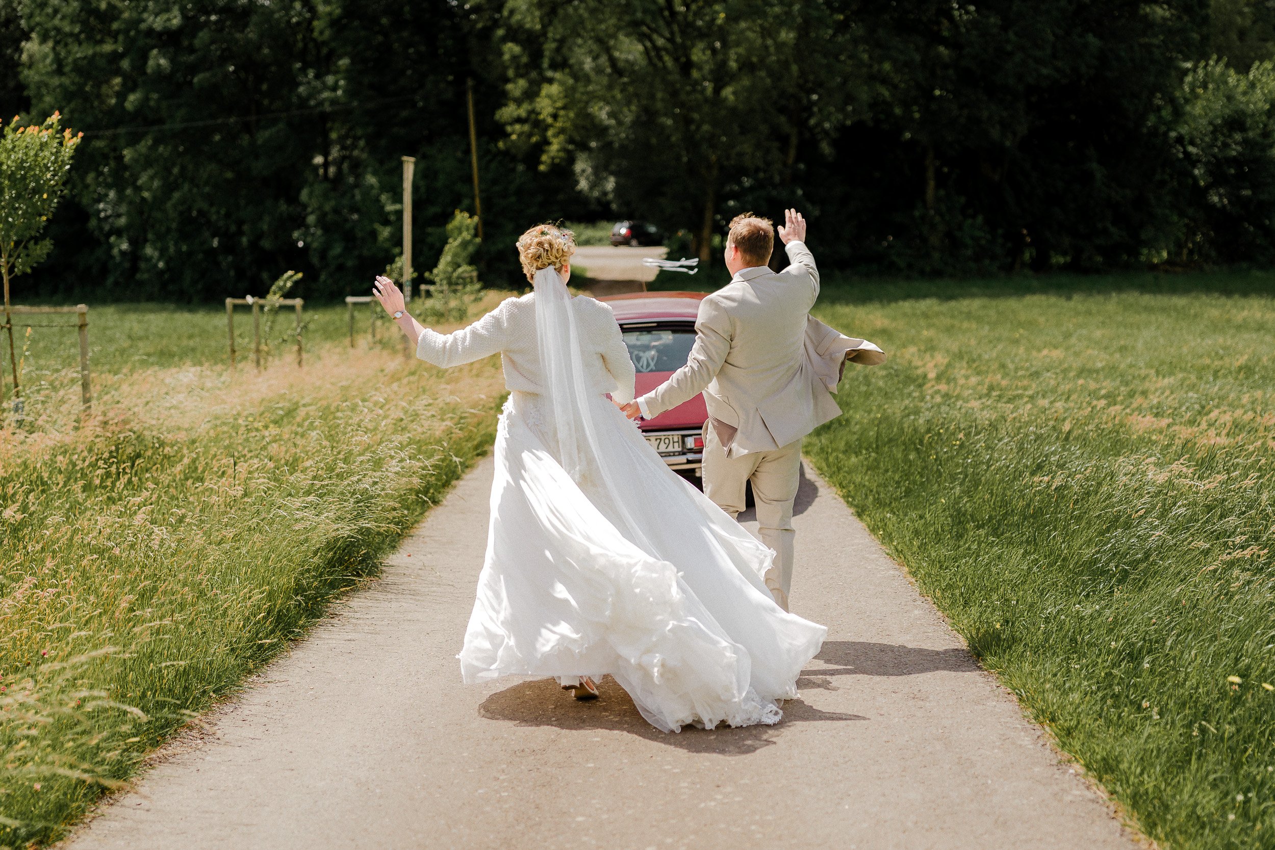 Ein frisch verheiratetes Paar läuft Hand in Hand auf einer Landstraße, umgeben von grünen Wiesen und Bäumen, an einem sonnigen Tag.