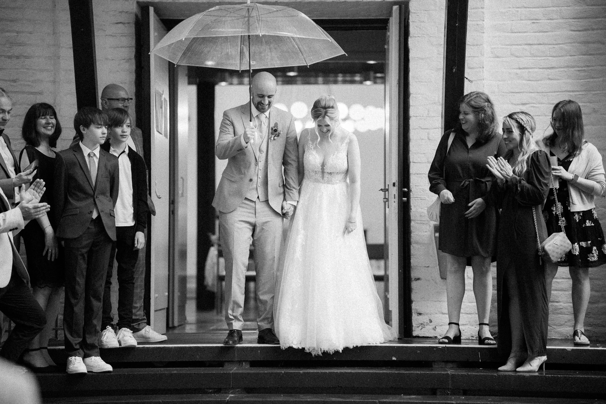 Braut und Bräutigam, die an ihrer Hochzeit Hand in Hand gehen, unter einem durchsichtigen Regenschirm, während Gäste ihnen applaudieren und zusehen.
