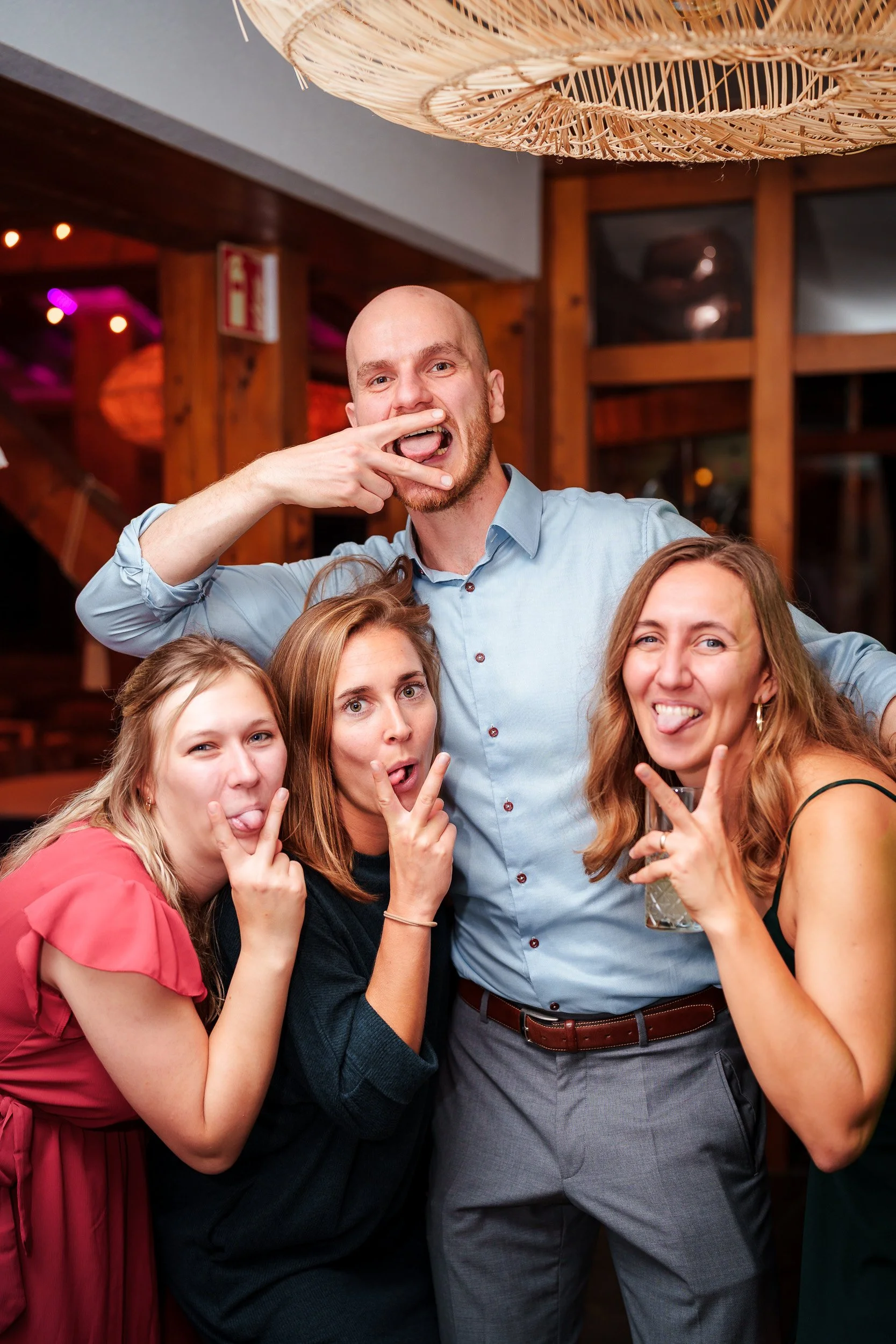 Vier junge Erwachsene feiern in einer Bar, machen Grimassen und posieren für das Foto, während eine bunte, freundliche Atmosphäre herrscht.