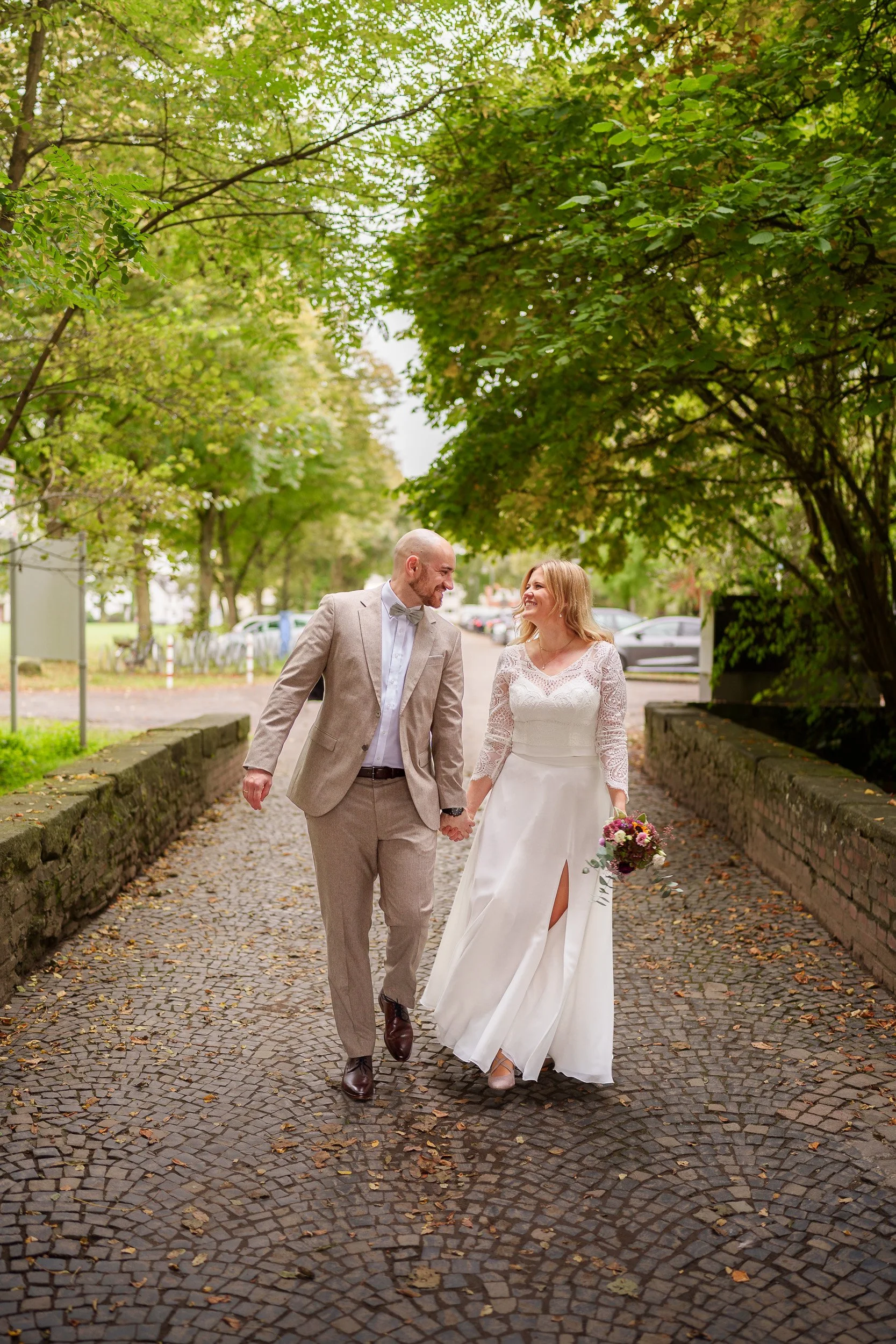 Ein Brautpaar geht Hand in Hand auf einer gepflasterten Straße, umgeben von grünen Bäumen, während es lächelnd und liebevoll in die Augen schaut.
