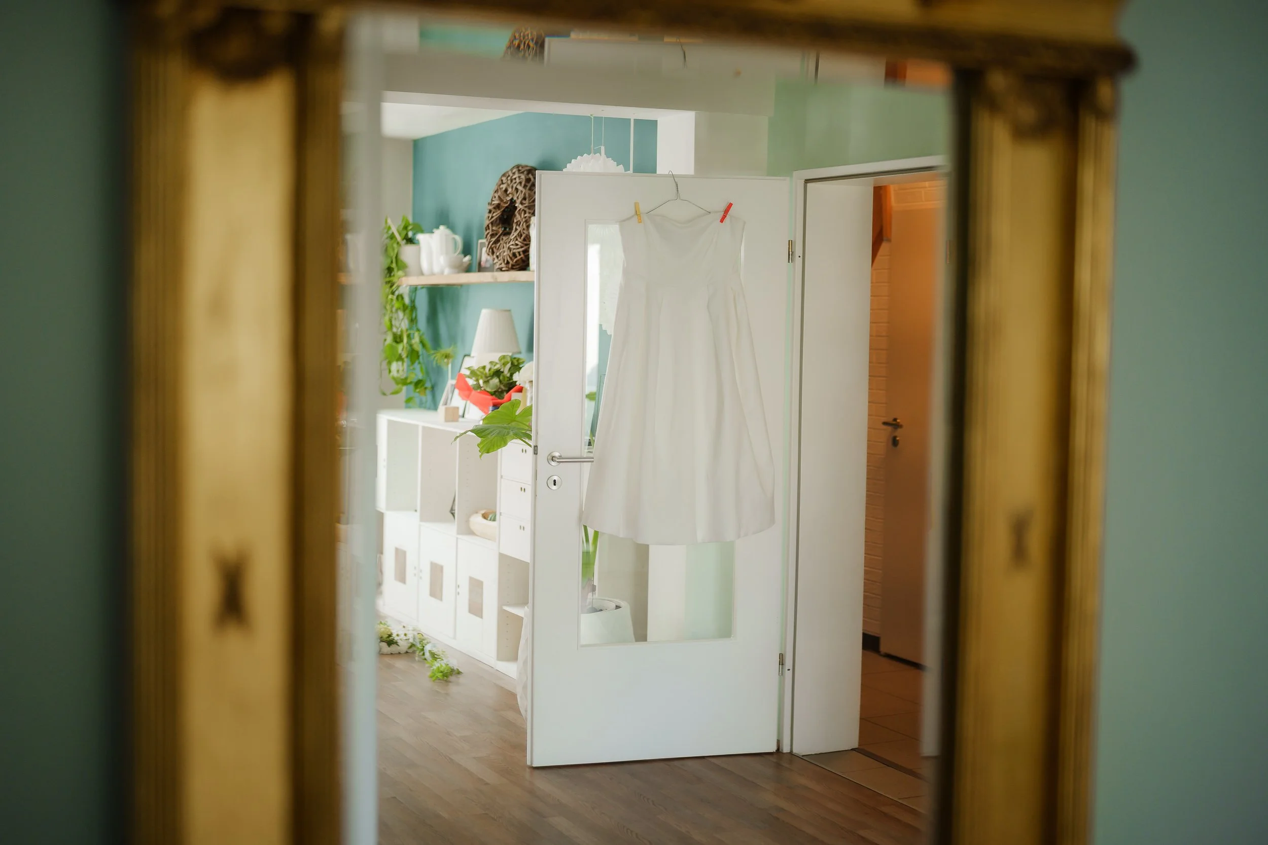 Hochzeitskleid hängt an einer Tür in einem hellen, modern eingerichteten Raum, sichtbar in einem Spiegel. Im Hintergrund befinden sich Dekorationsgegenstände und Pflanzen.
