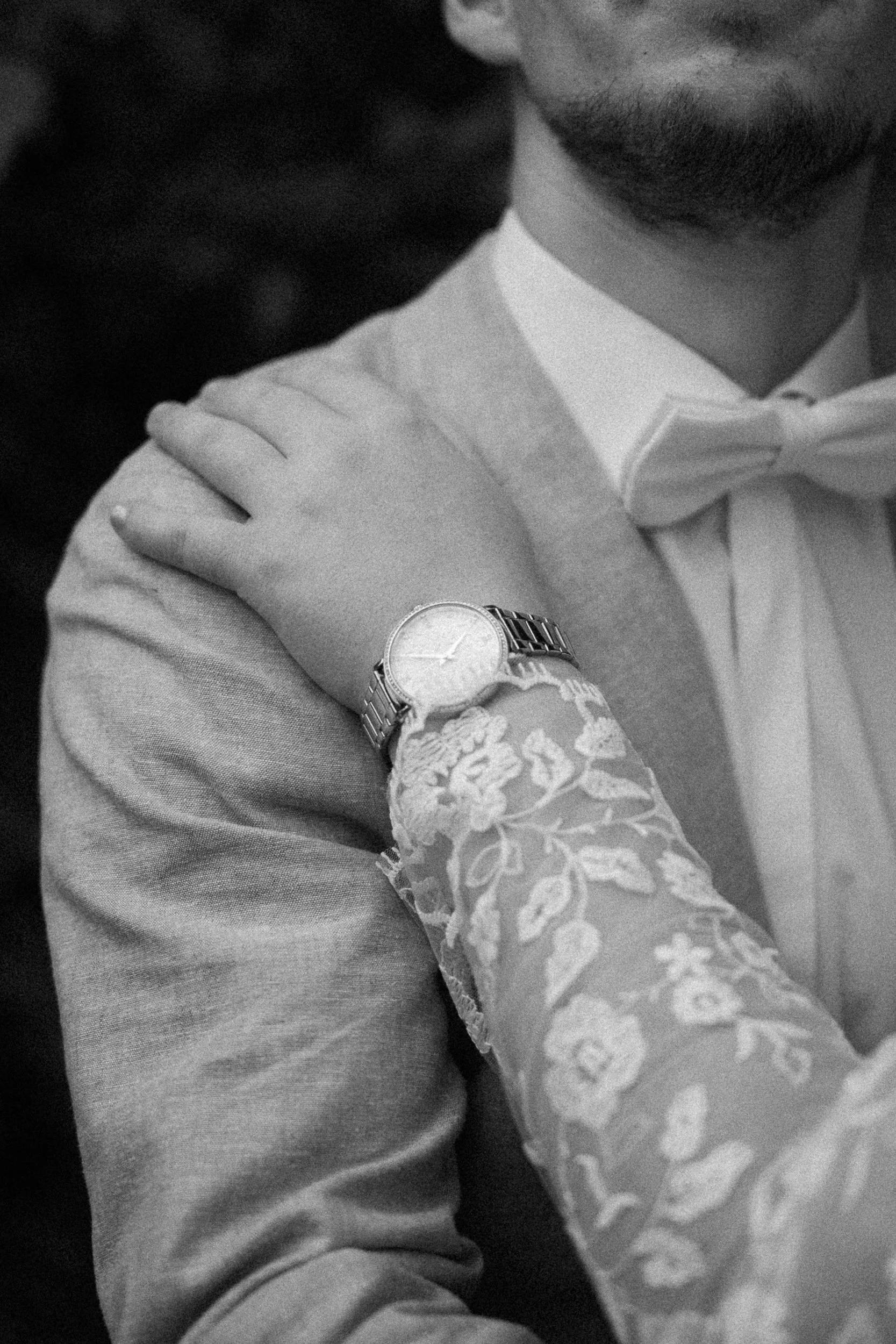 Ein Mann in formeller Kleidung mit Krawatte, trägt eine Armbanduhr. Eine Hand mit einer langen, gemusterten Spitze berührt seine Schulter.
