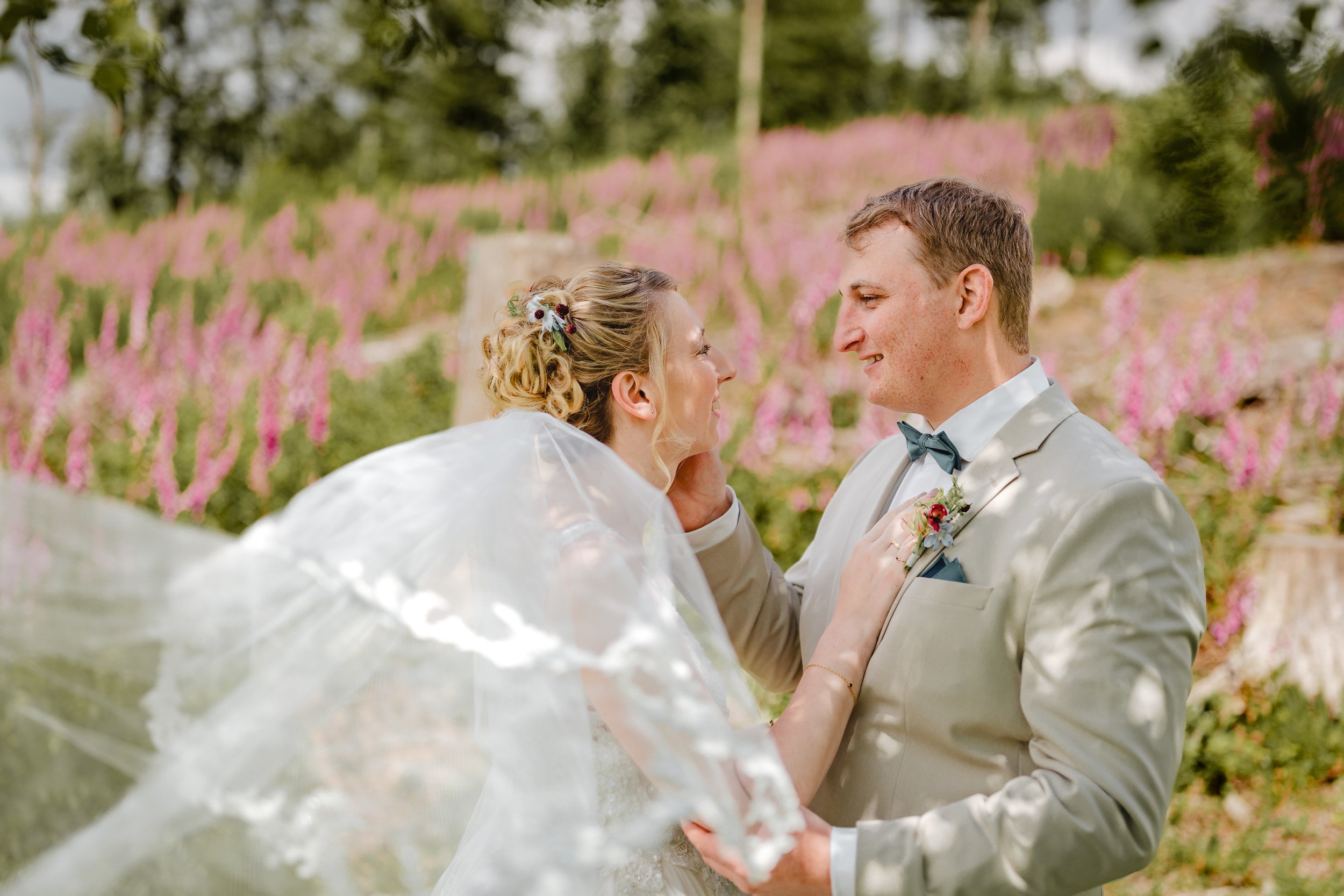 Ein Brautpaar steht sich bei einer Hochzeit im Freien gegenüber, umgeben von pinken Blumen und grünen Bäumen.