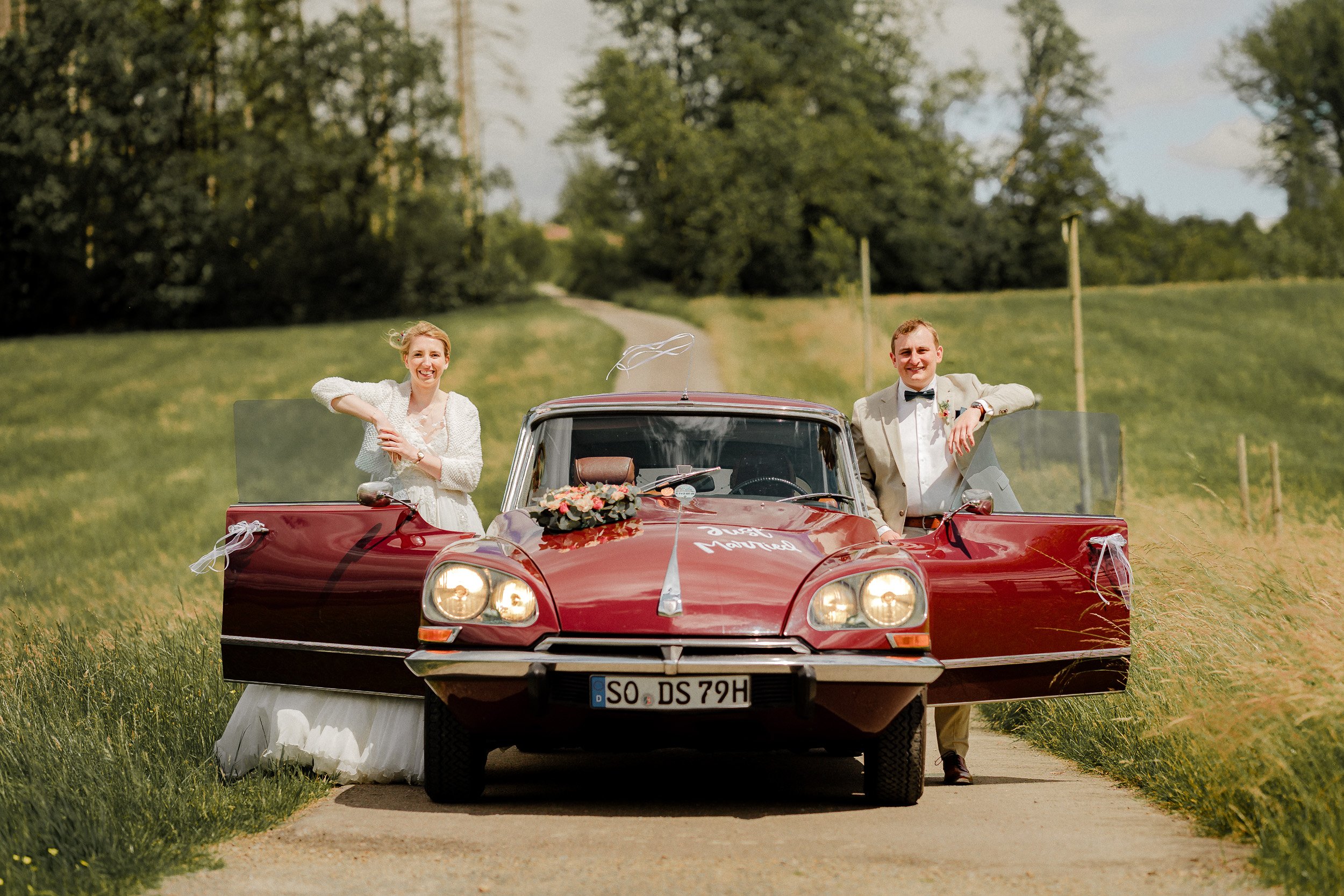 Ein Paar feiert eine Hochzeit mit einem roten Oldtimer in einer ländlichen Gegend, grüne Wiesen und Bäume im Hintergrund.