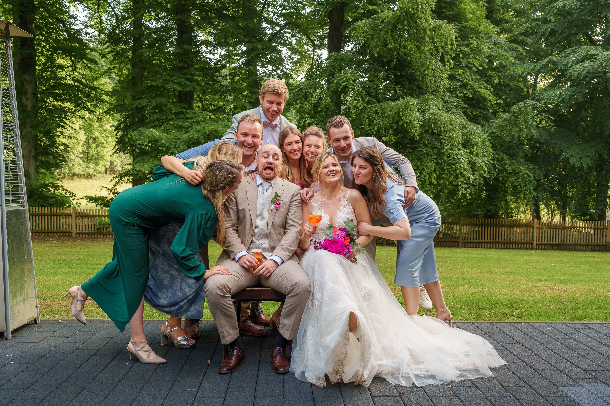 Gruppenfoto von Hochzeitsgesellschaft im Grünen, mit Braut und Bräutigam in der Mitte, umgeben von Freunden, alles lächelt und hat Spaß.