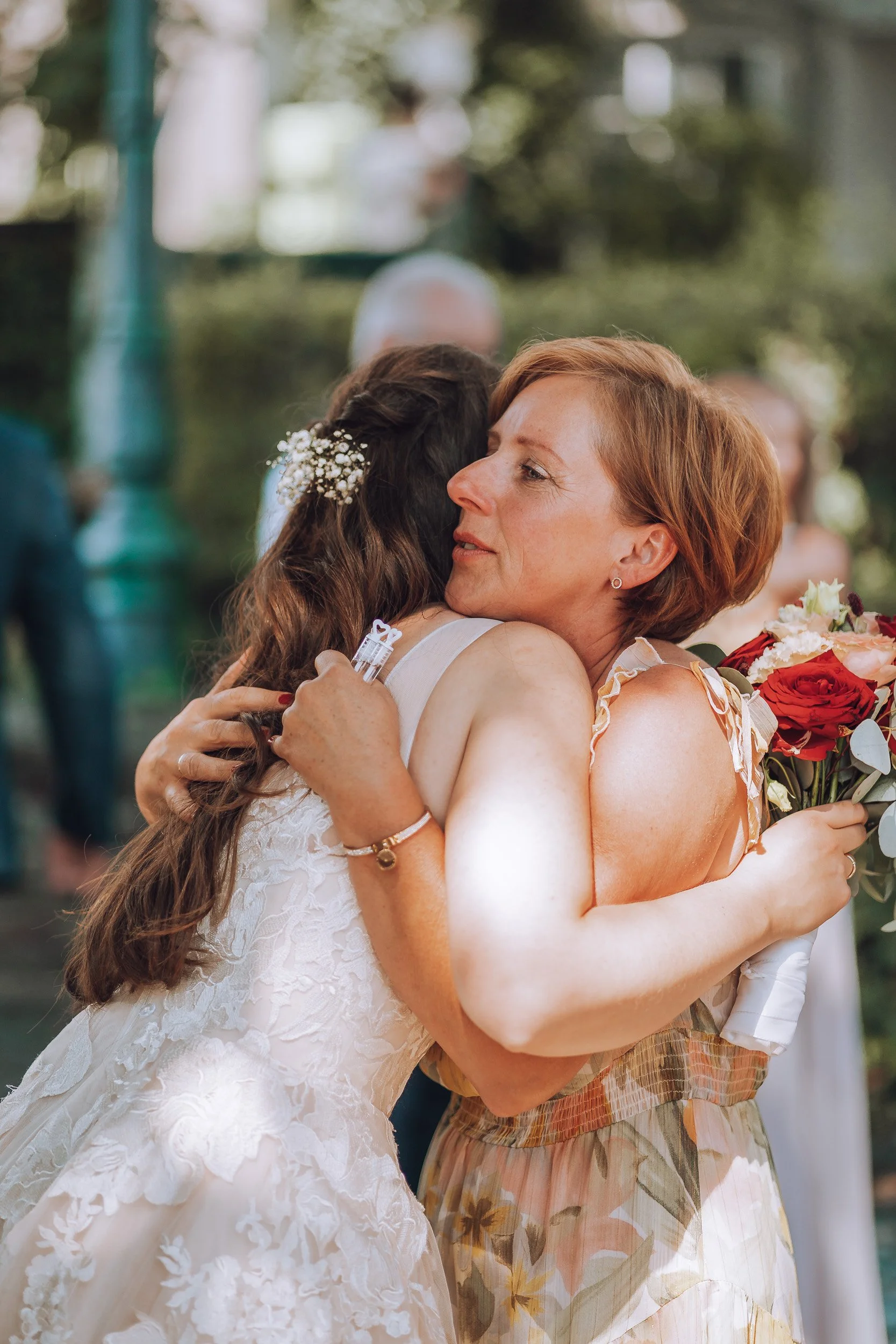 Zwei Frauen umarmen sich bei einer Hochzeit, eine trägt ein Brautkleid, die andere hält einen Blumenstrauß, in einem Garten.