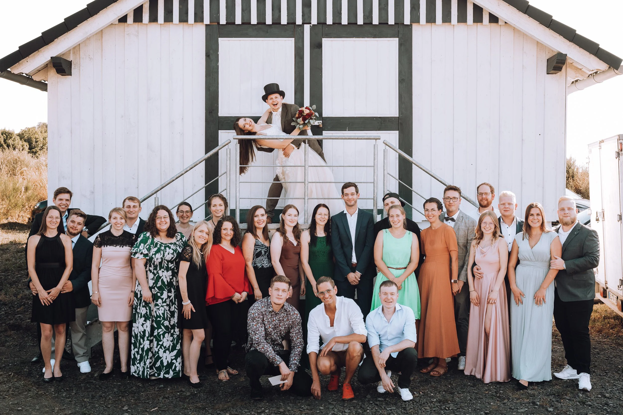 Gruppenfoto bei einer Hochzeit vor einem weißen Holzgebäude mit schwarzen Rahmen, mit Brautpaar oben auf der Treppe, der Bräutigam trägt einen Zylinderhut, die Braut hält einen Blumenstrauß, umgeben von Freunden und Familie.