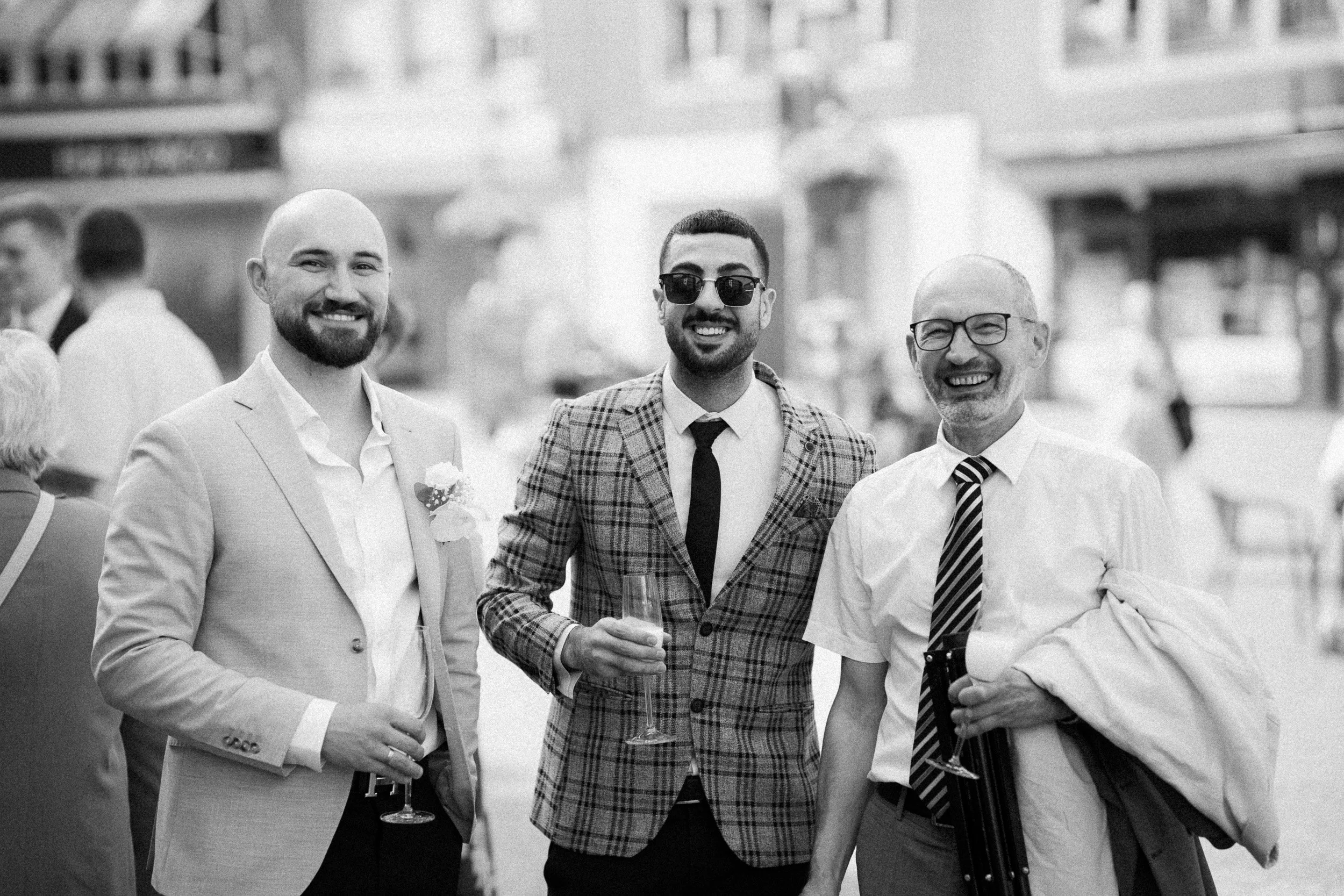 Drei Männer bei einer Feier, alle lächeln und halten Gläser mit Getränken, im Hintergrund sind andere Gäste und eine Stadtumgebung sichtbar.