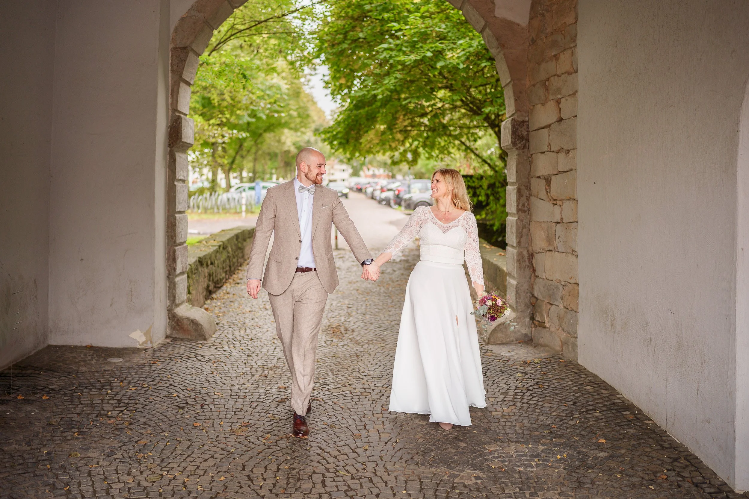 Ein glückliches Paar in Hochzeitskleidung, Hand in Hand, unter einer Steinbrücke, umgeben von grünen Bäumen und einem gepflasterten Weg.