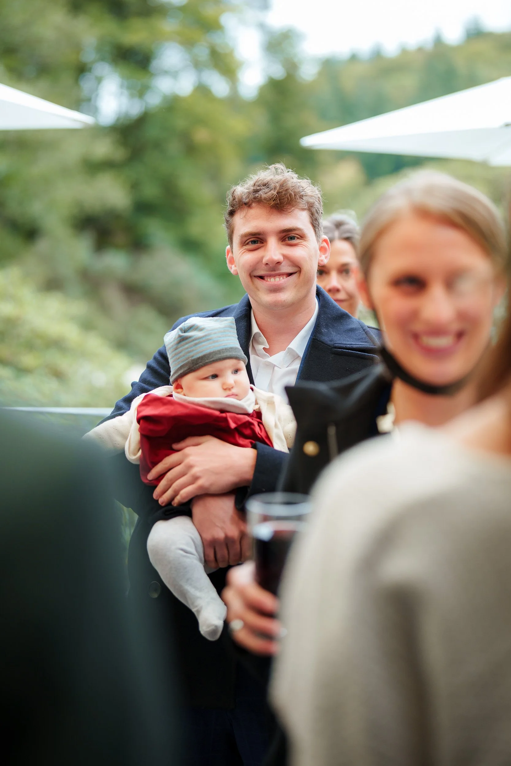 Menschen bei einer Zusammenkunft im Freien, im Hintergrund ein lachender junger Mann, ein Baby in den Armen eines Mannes, umgeben von mehreren lachenden Menschen, im Vordergrund eine lachende Frau mit einem Glas Rotwein.