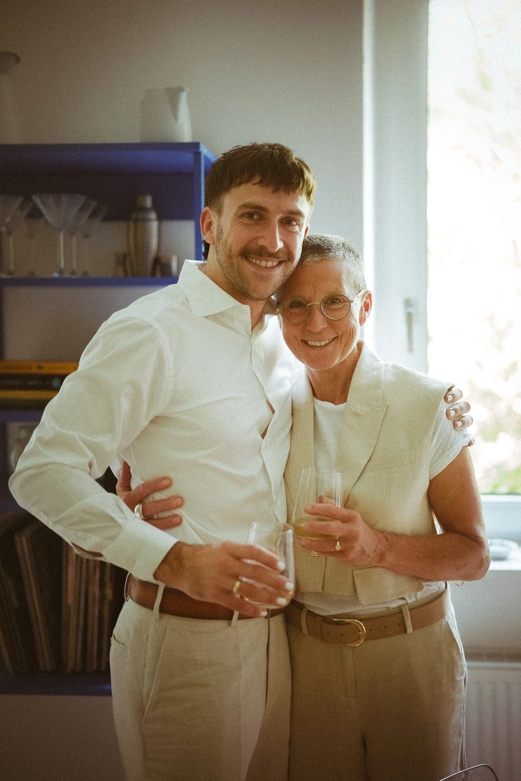 Zwei Menschen, ein Mann und eine Frau, die beide ein Glas Wein halten, umarmen sich lächelnd in einem Raum mit natürlichem Licht.
