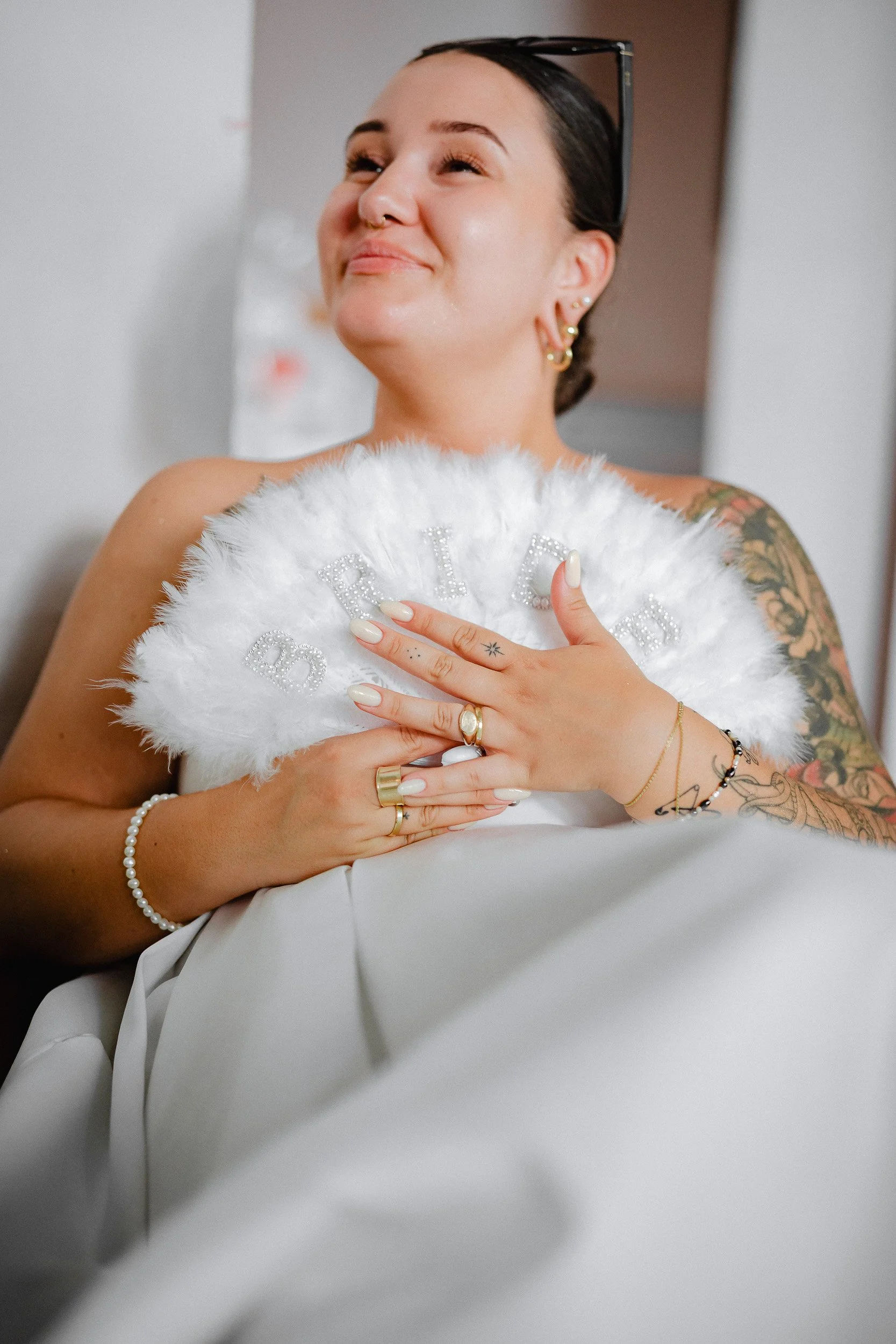 Eine strahlende Frau mit Tattoos, Schmuck und Brille auf dem Kopf hält eine weiße, pelzige Dekoration mit den Buchstaben 'BRIDE' in der Hand vor ihrer Brust, lächelt zufrieden.