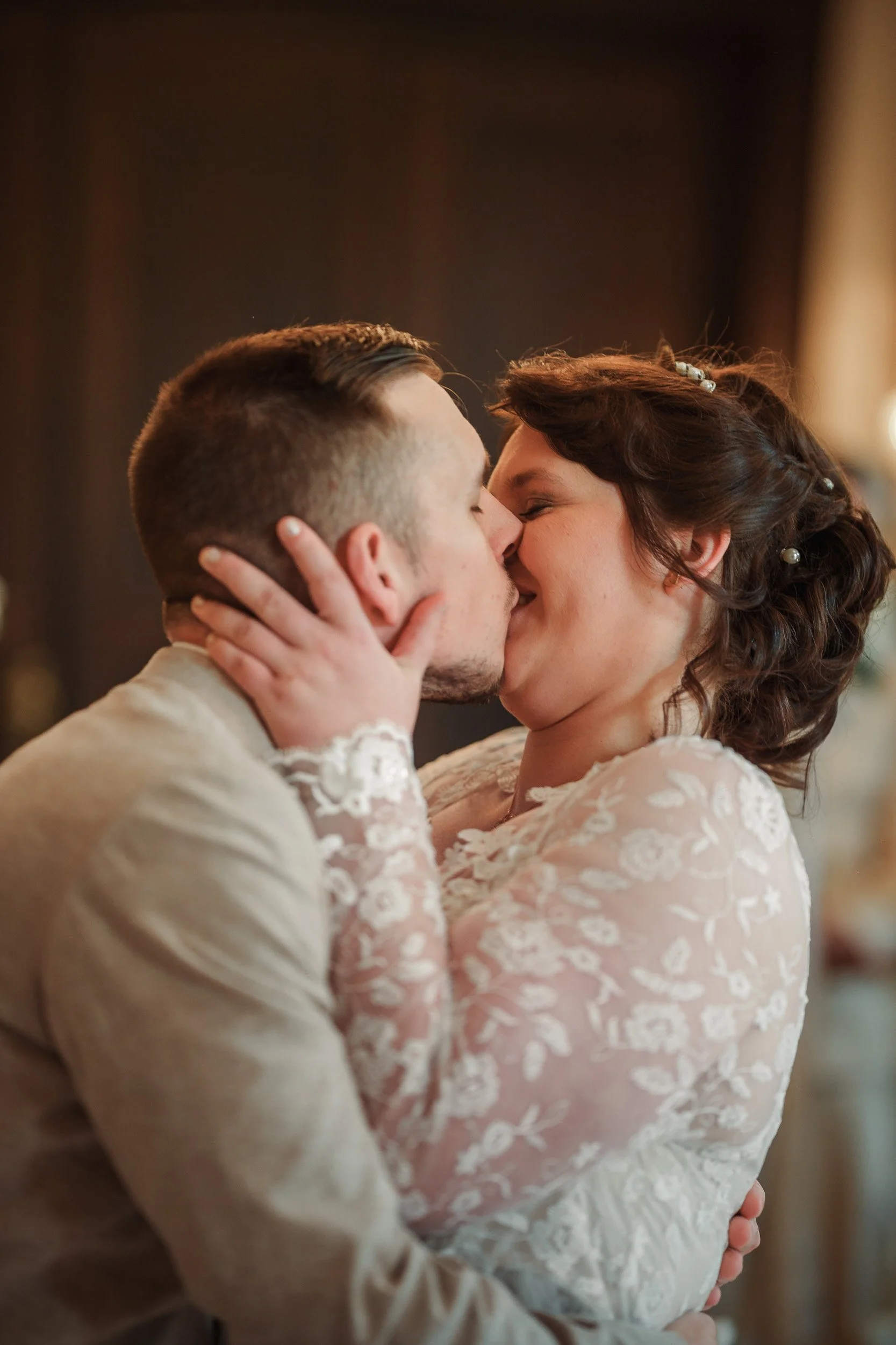 Ein verliebtes Paar küsst sich bei einer Hochzeit, die Frau trägt ein weißes Spitzenkleid, der Mann ein blasses Jackett.