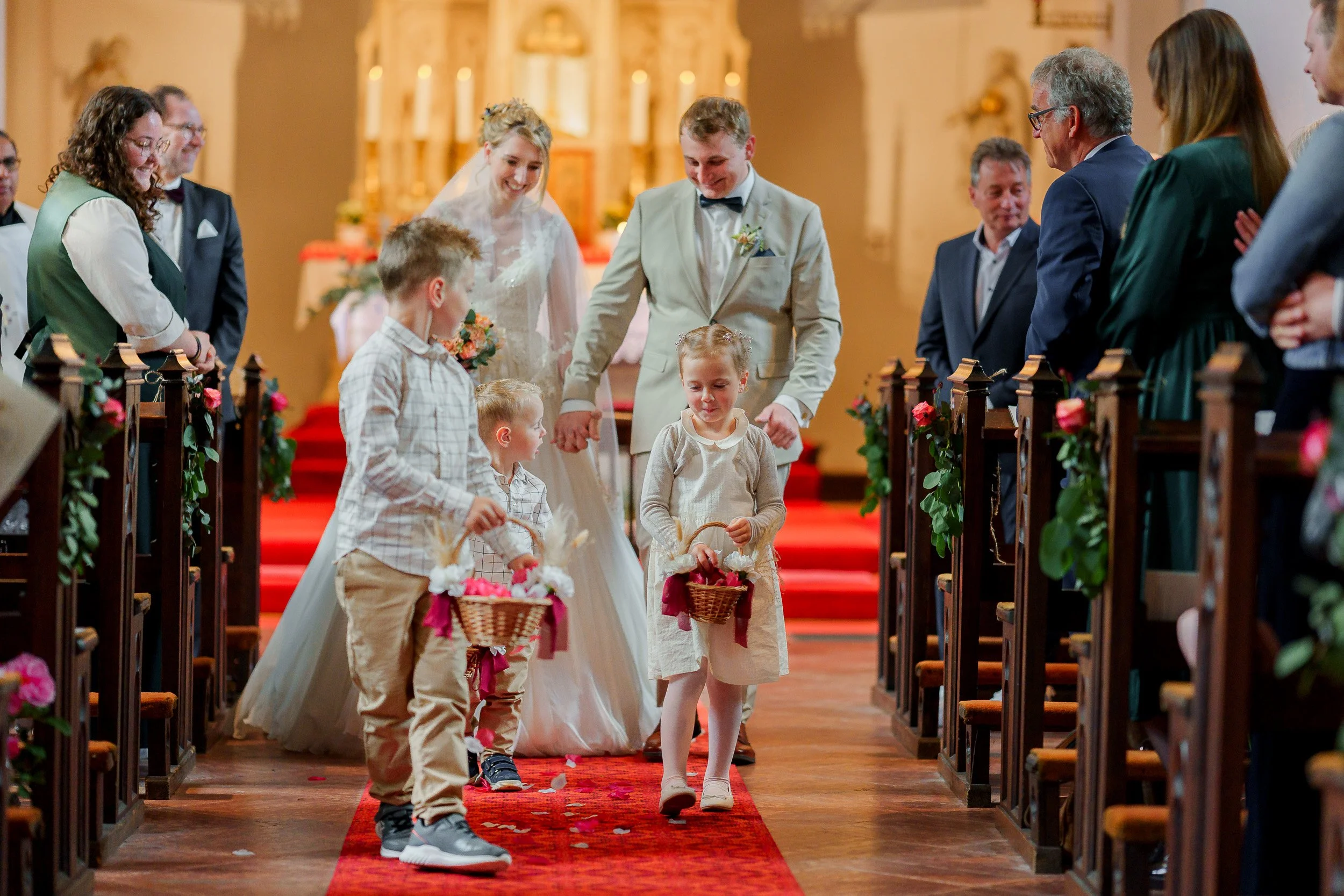 Hochzeitszeremonie in einer Kirche mit Braut, Bräutigam, Gästeschar und Kindern, die beim Einzug Blumen und Körbe mit Blumen streuen.