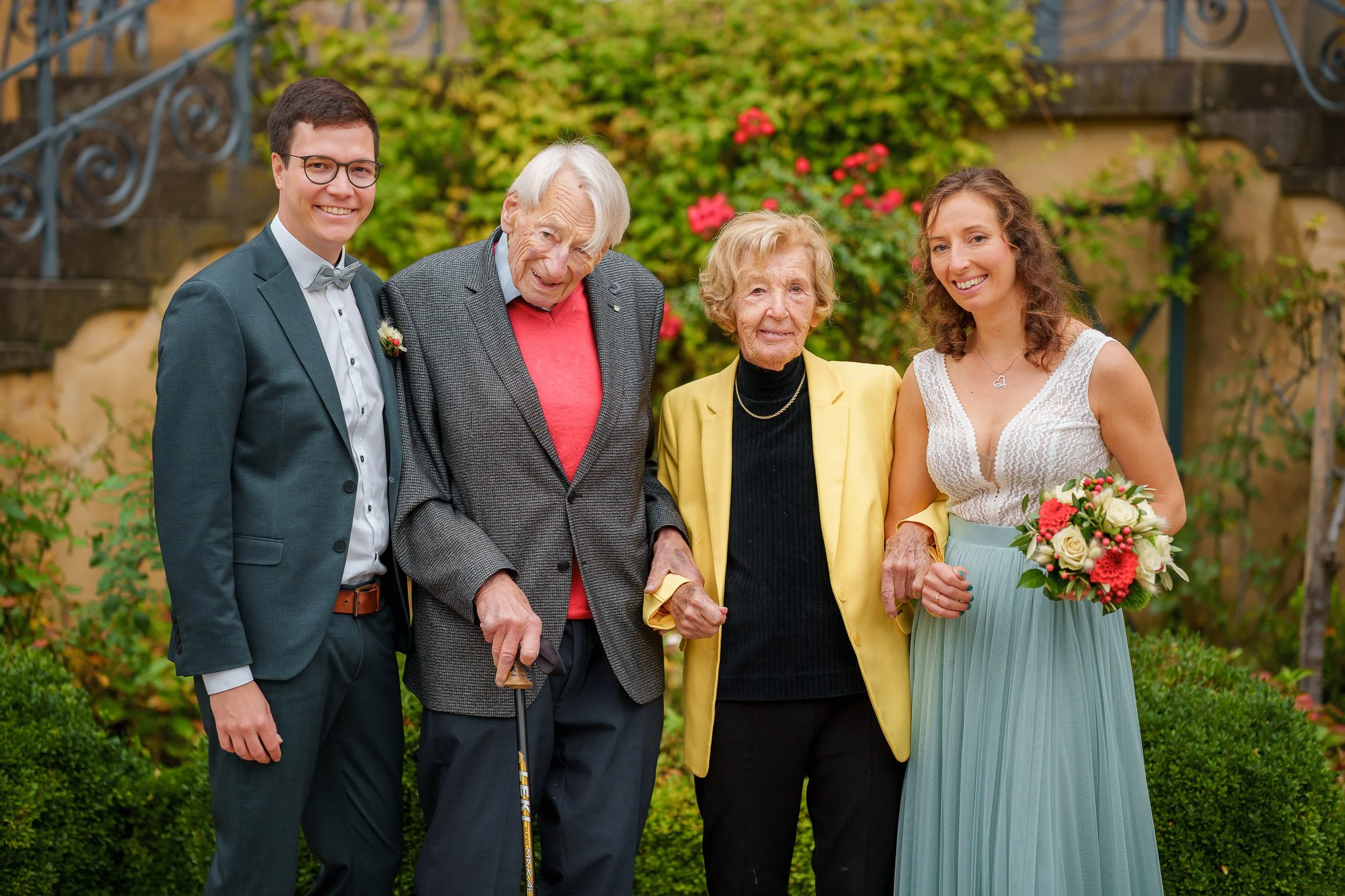 Familienfoto mit fünf Personen bei einer Hochzeit, inklusive älterer Männer und Frauen, sowie jüngere Frauen, im Garten mit Blumen. Der Mann auf der linken trägt einen Anzug, die ältere Frau in der Mitte trägt ein schwarzes Oberteil und eine gelbe Ja