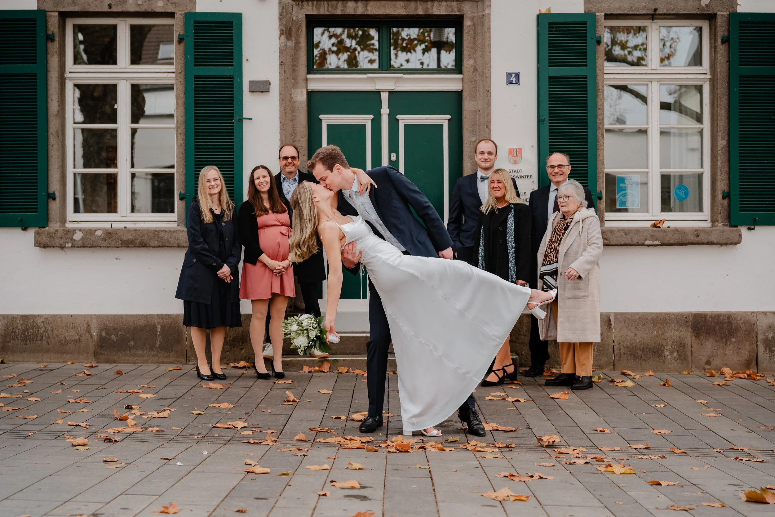 Ein frisch vermähltes Paar teilt sich einen Kuss vor einem historischen Gebäude, umgeben von Familienmitgliedern und Freunden, im Freien bei herbstlichem Wetter mit Blättern auf dem Boden.