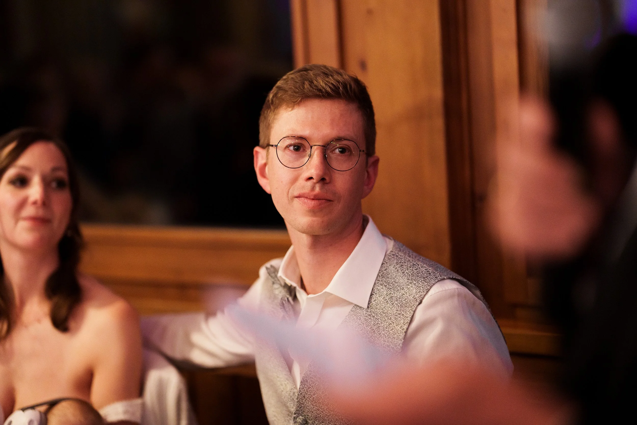 Junger Mann mit Brille in einem festlichen Raum, im Gespräch mit anderen Menschen.