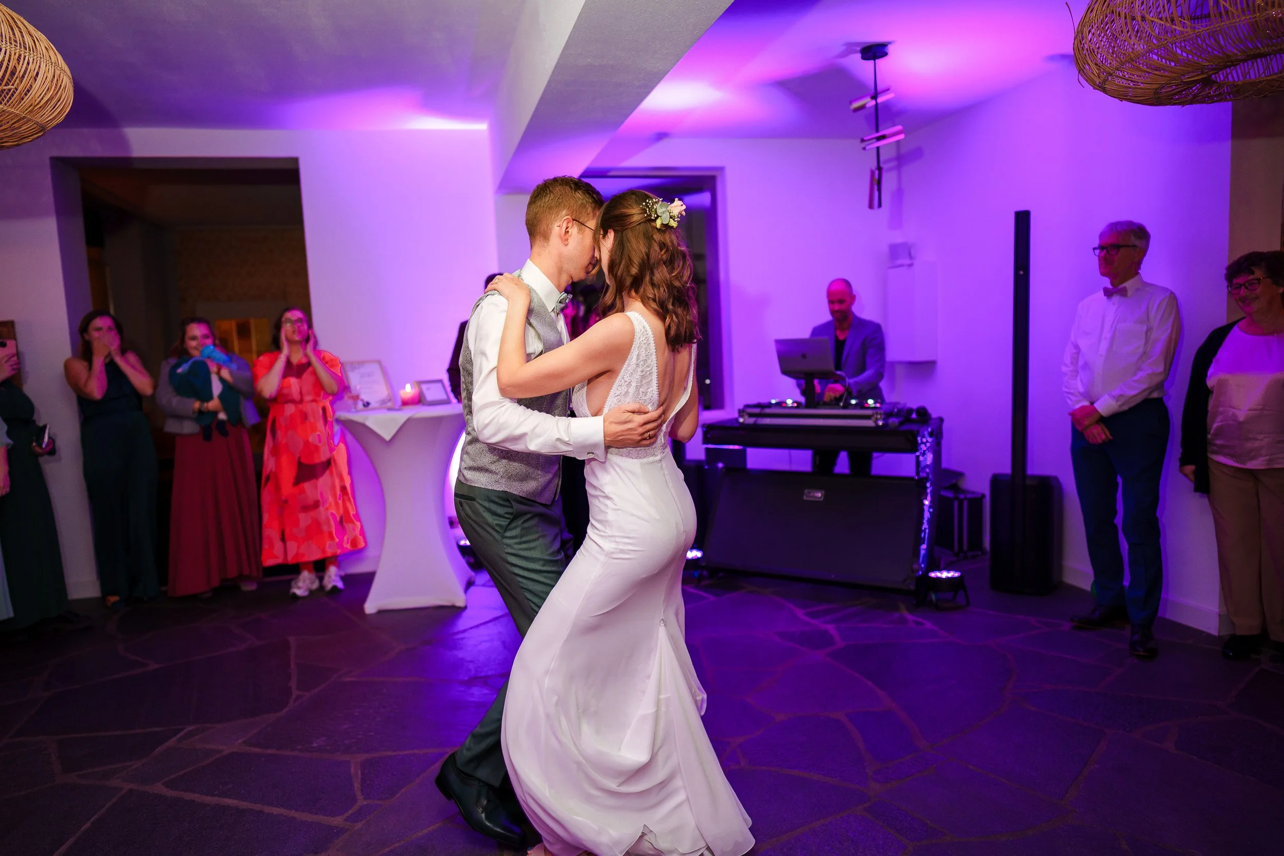 Braut und Bräutigam tanzen auf der Hochzeit, umgeben von Gästen, im Hintergrund ein DJ und bunte Lichter.