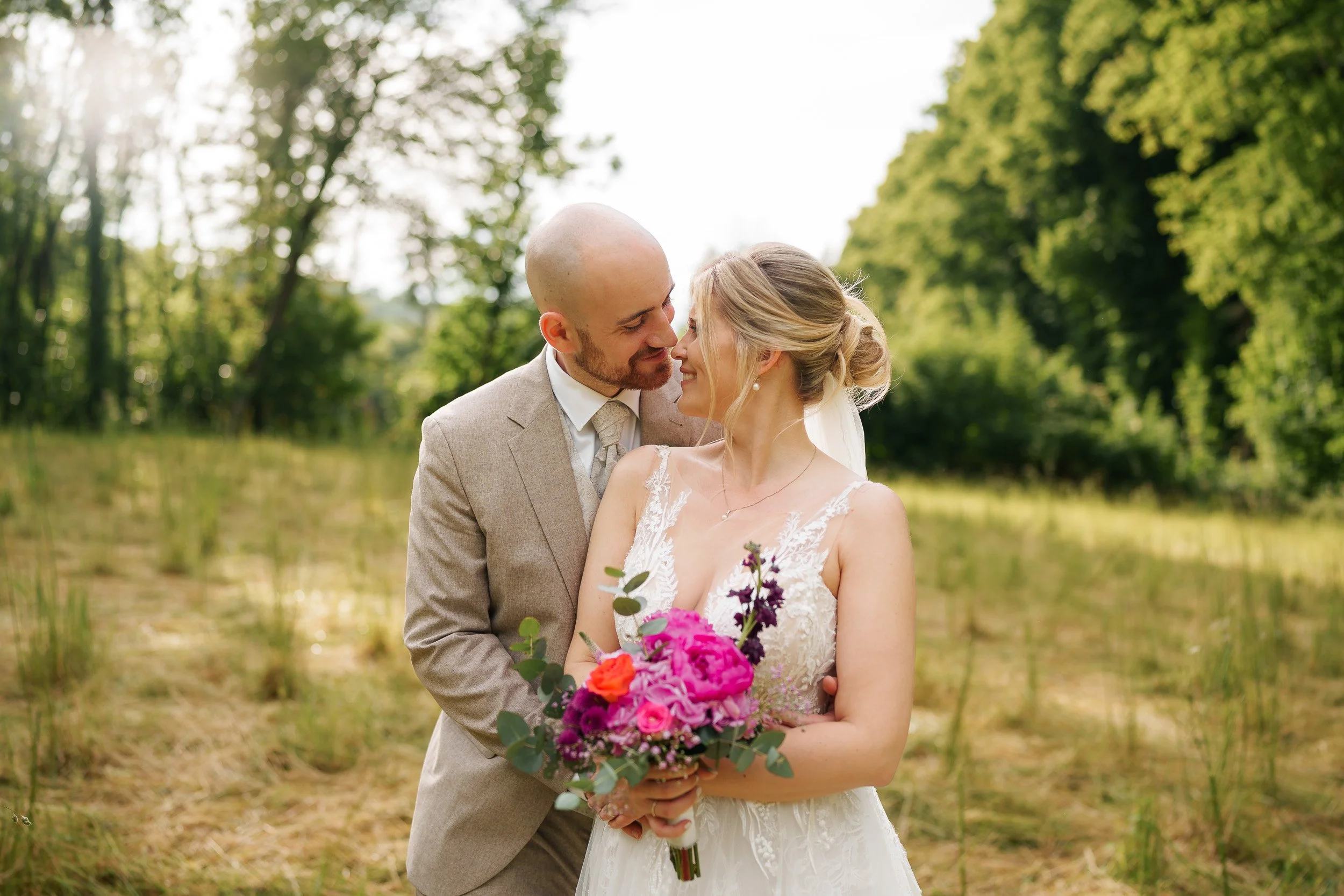 Ein lächelndes Brautpaar steht im Grünen, die Braut hält einen Blumenstrauß, sie schauen sich liebevoll an.