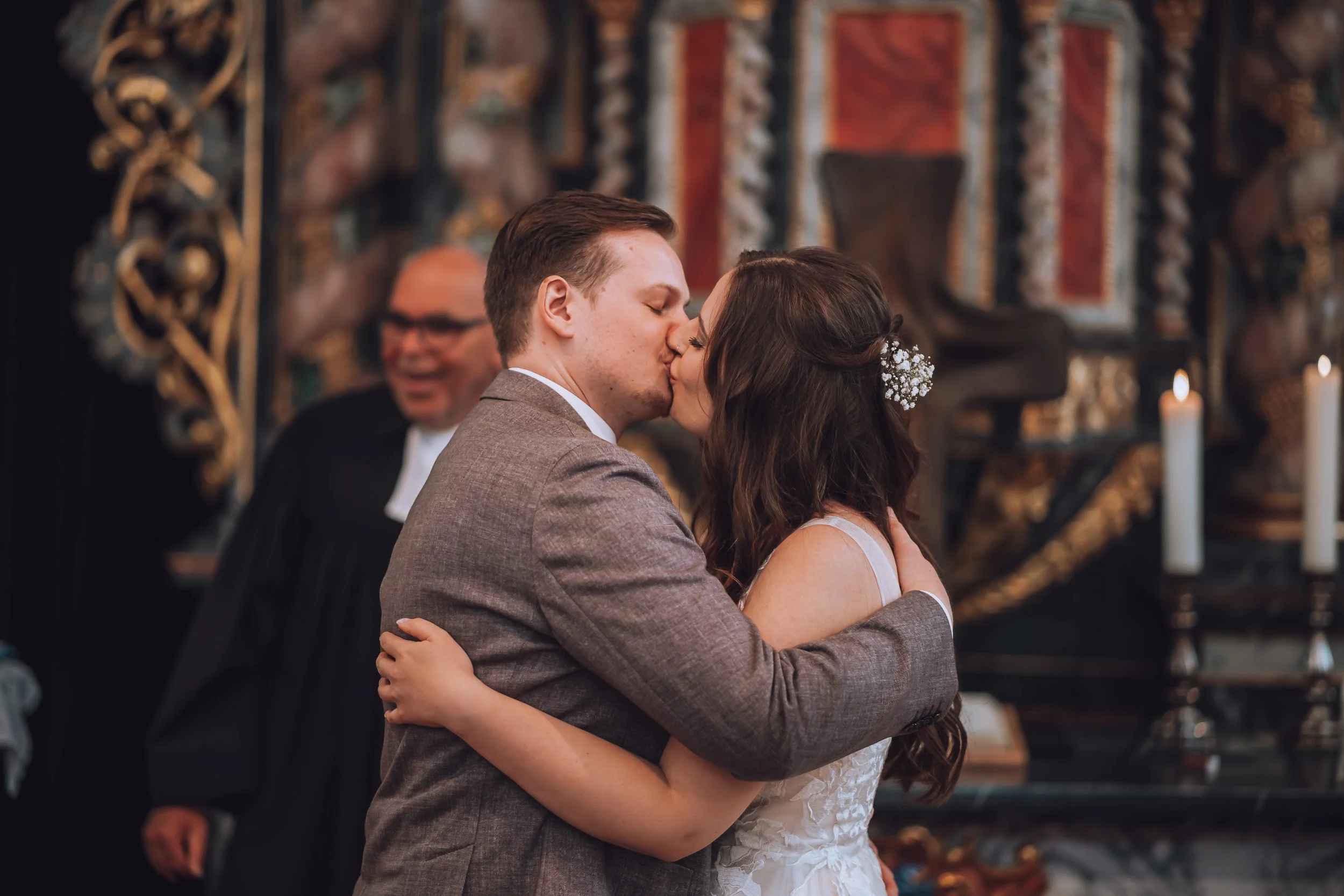 Ein Brautpaar küsst sich während einer Hochzeit, im Hintergrund jemand, der als Offizieller oder Geistlicher gekleidet ist, in einer Kirche oder einem Festsaal mit Kerzen und kunstvollen Dekorationen.