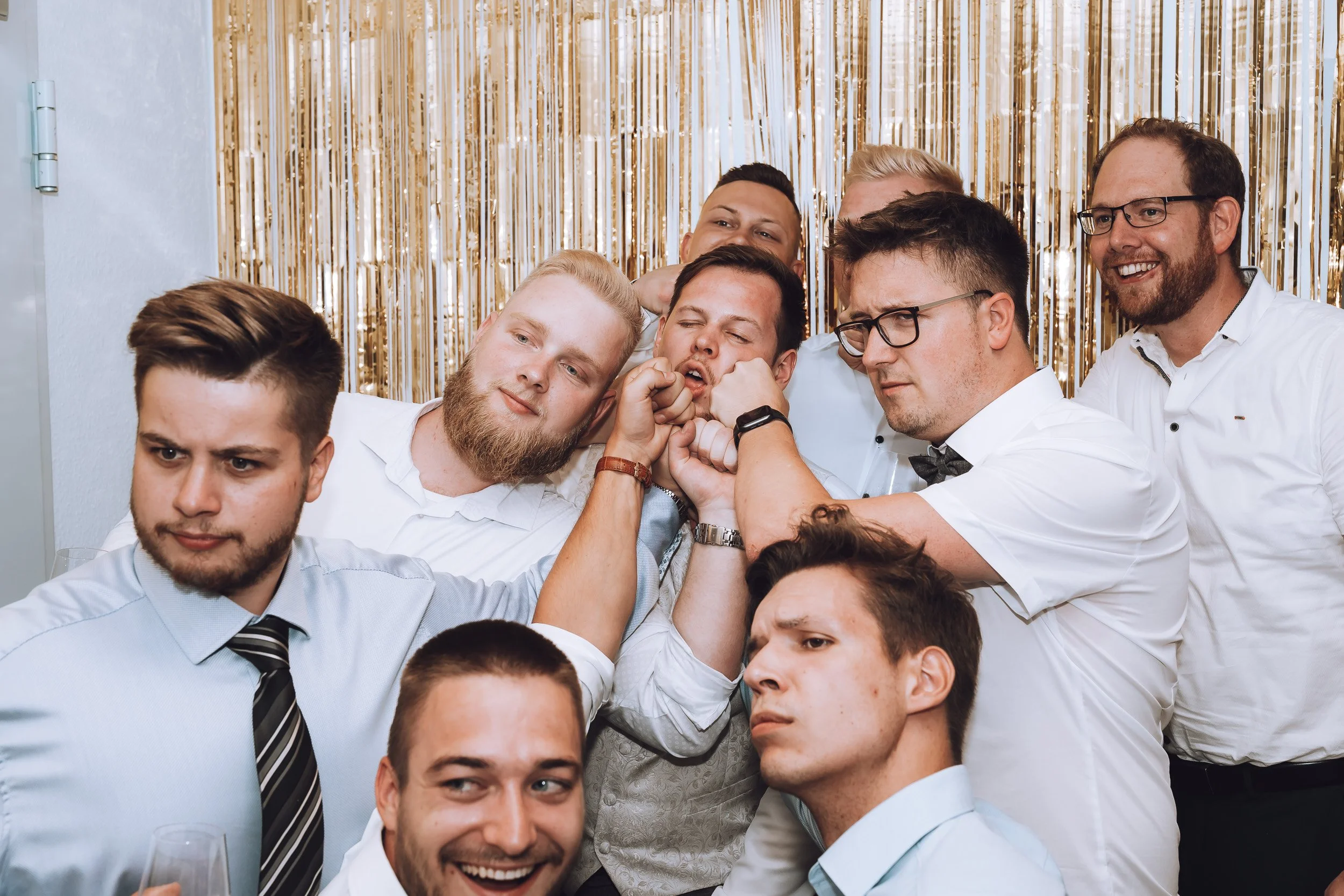 Gruppe junger Männer beim Kampf beim Feiern, in formeller Kleidung, mit goldener Dekoration im Hintergrund.