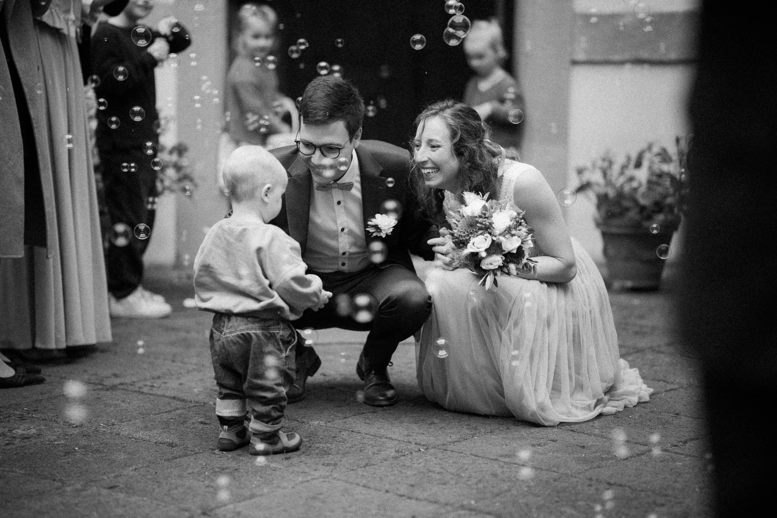 Ein Brautpaar und ein kleiner Junge bei einer Hochzeit, umgeben von Seifenblasen und Gästen im Hintergrund.
