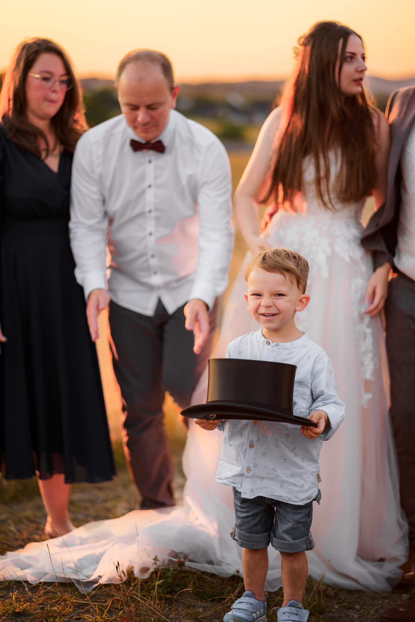 Ein Junge hält einen schwarzen Zylinderhut und lächelt bei einem Outdoor-Event bei Sonnenuntergang, umgeben von Erwachsenen in festlicher Kleidung.