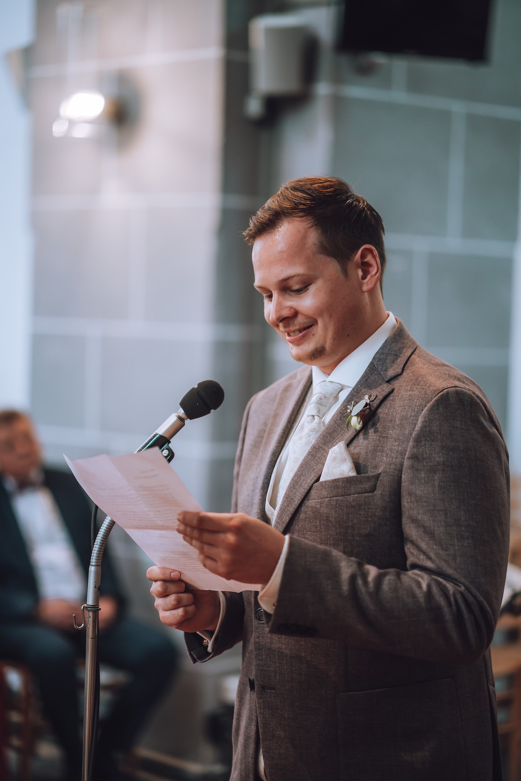 Der Bräutigam liest eine Rede während einer Hochzeitsfeier, trägt einen braunen Anzug und steht vor einem Mikrofon.