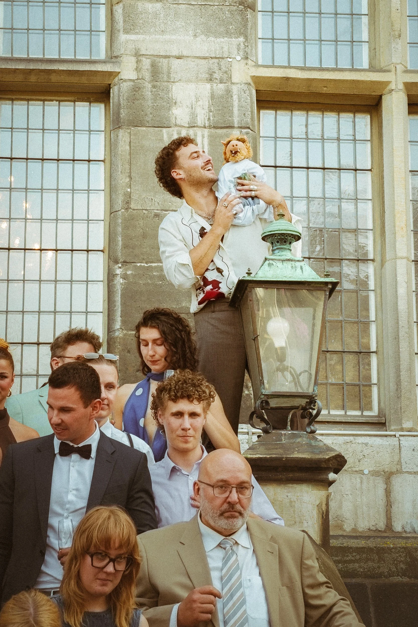 Menschen versammeln sich vor einem alten Gebäude mit großen Fenstern. Ein Mann hält eine Pampe mit einem Puppenkopf eines Löwen an der Hand und scheint lacht. Es ist eine Gruppe von Menschen, die sich in einer sozialen oder öffentlichen Veranstaltung