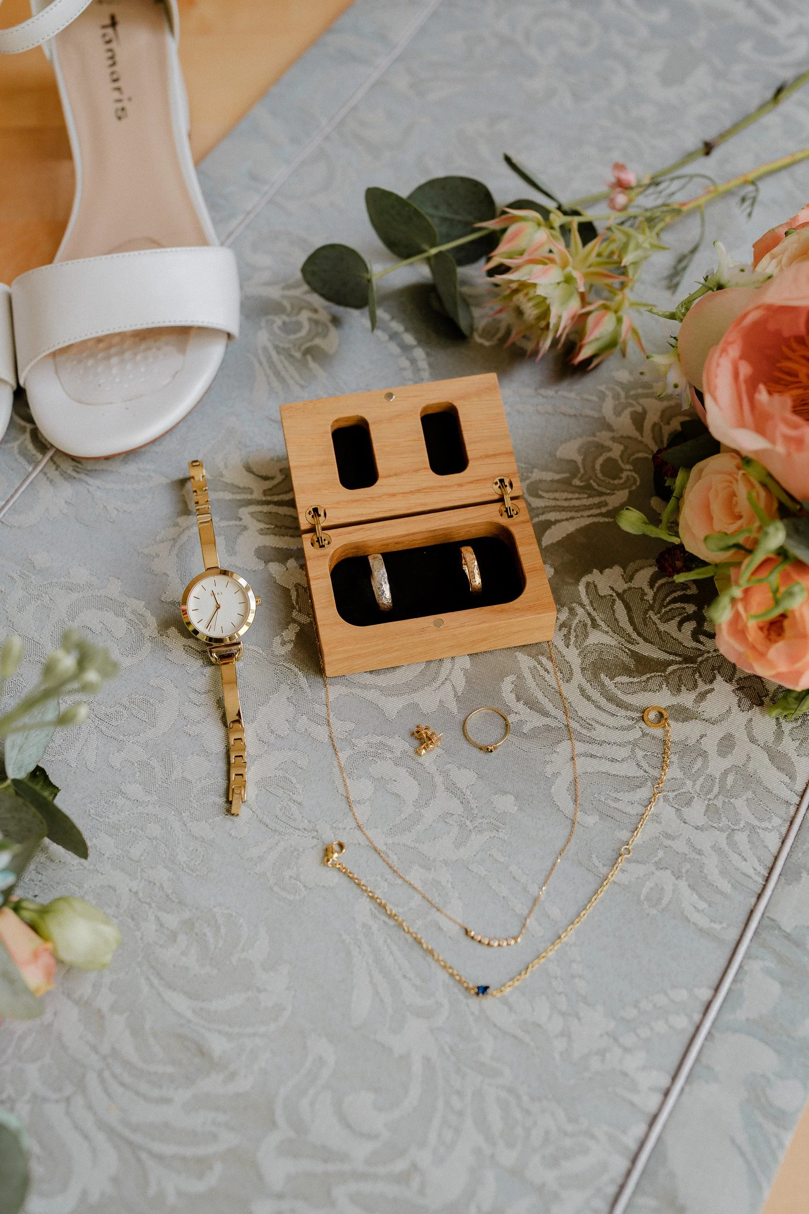 Ringe | Schmuck und Schuhe für eine Hochzeit, darunter ein goldener Armreif, Ringe, eine Halskette, Ohrringe, ein Schuh, ein Holzschmuckkästchen, Blumen und eine Tischdecke.
