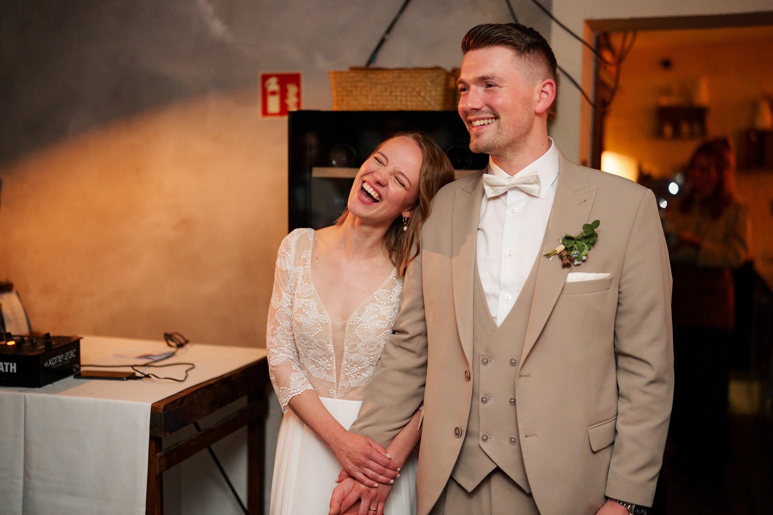 Lächelndes Brautpaar bei einer Hochzeitsfeier, die Braut trägt ein weißes Kleid mit Spitzenüberwurf, der Bräutigam trägt einen beigen Anzug mit Krawatte und Blumenschmuck auf der Jacke, während sie Händchen halten.