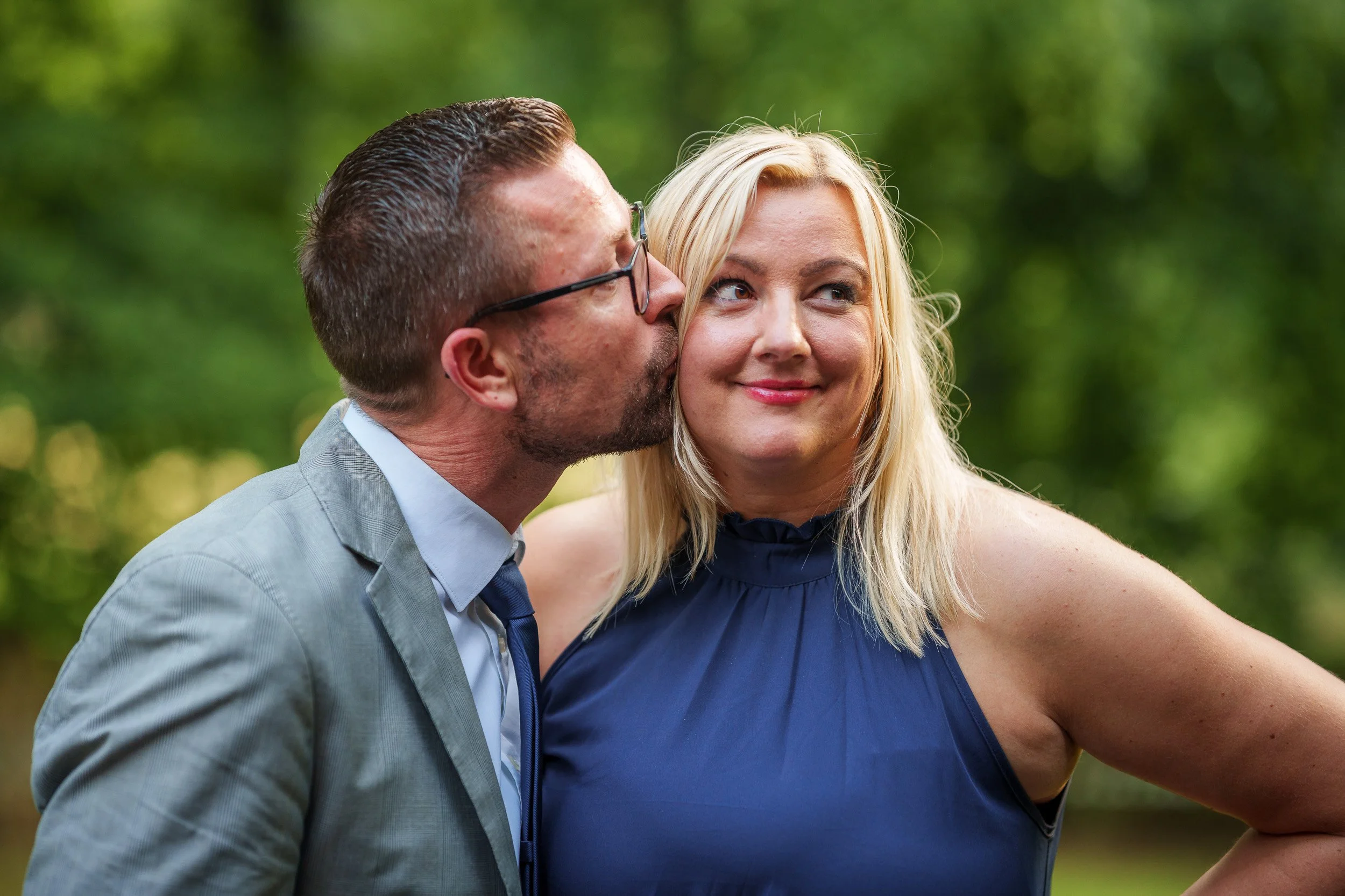 Ein Mann in Brille küsst eine lachende Frau auf die Wange in einem Park.