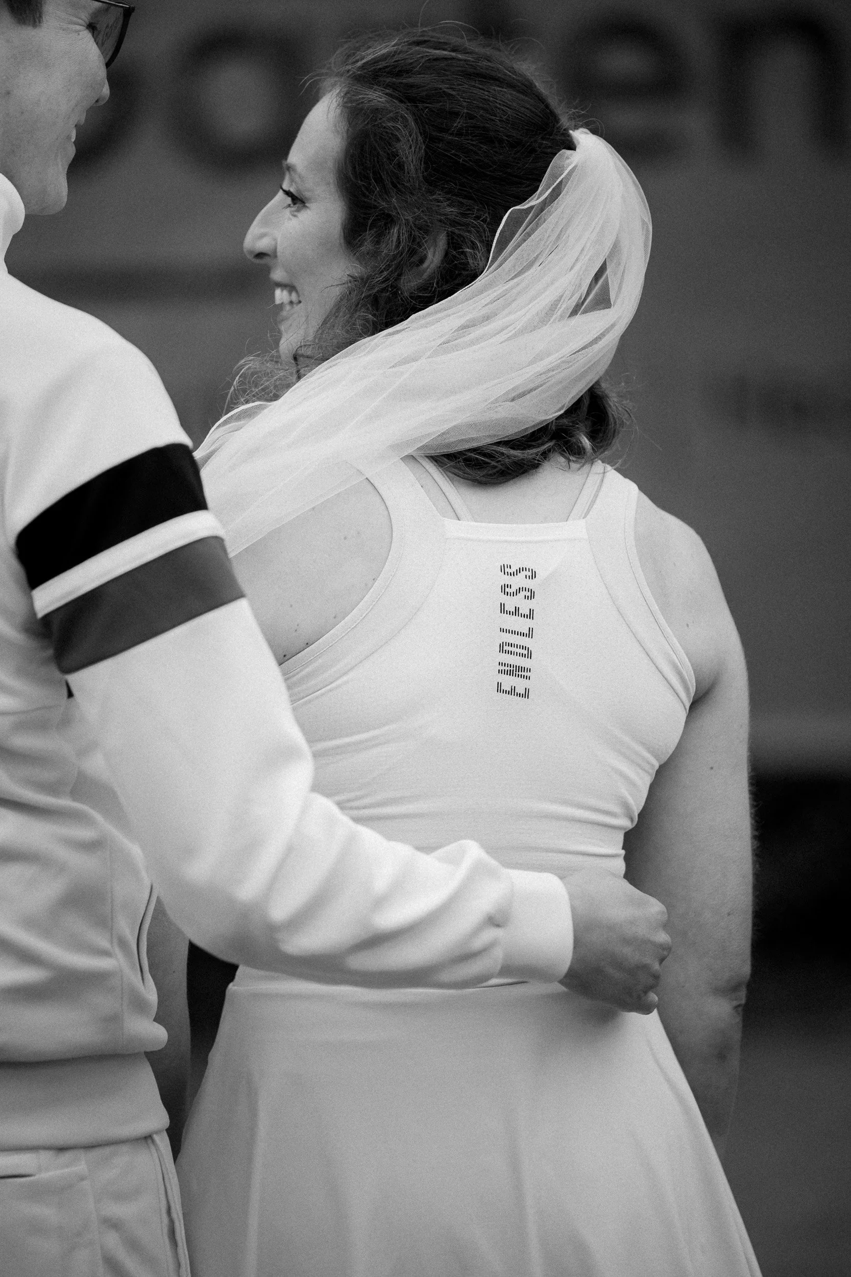 Eine Frau trägt ein Hochzeitskleid und einen Schleier und lächelt, während sie mit einem Mann spricht, der aus einer Sportjacke besteht. Das Foto ist schwarz-weiß.