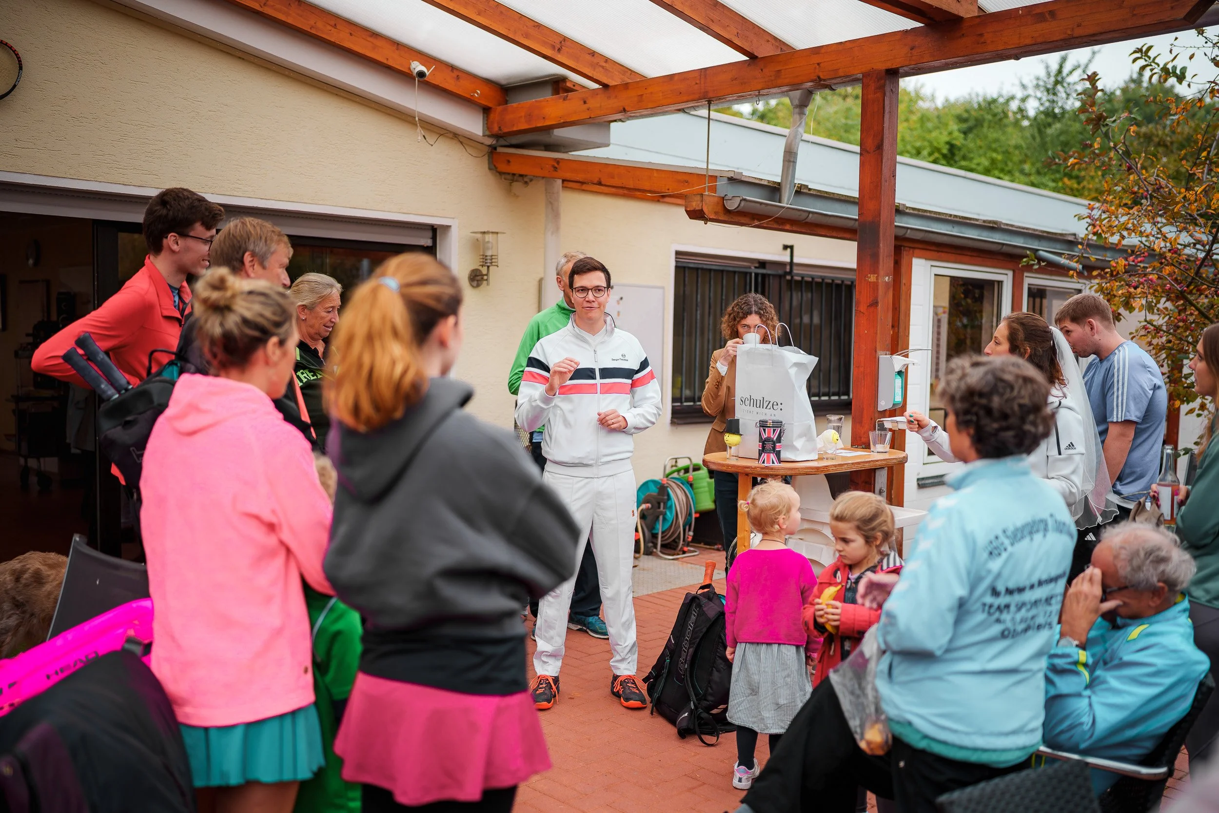 Eine Gruppe von Menschen, darunter Kinder und Erwachsene, versammelt sich auf einer Terrasse im Freien bei einem Treffen oder einer Feier, mit einem Mann, der vermutlich eine Rede hält.